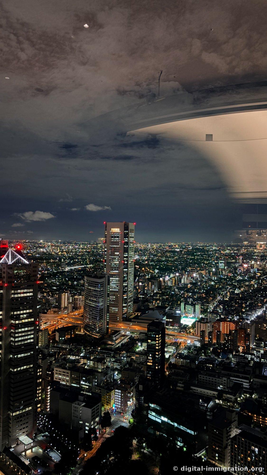 Vue nocturne en direction d'Opera City à travers une vitre reflétant grandement l'éclairage du plafond