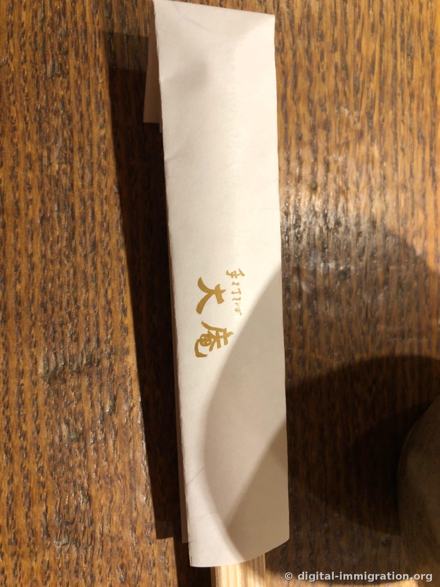 나무 테이블 위에 놓인 흰 젓가락 봉투, 금색으로 手打ちそば 大庵이라고 인쇄되어 있다