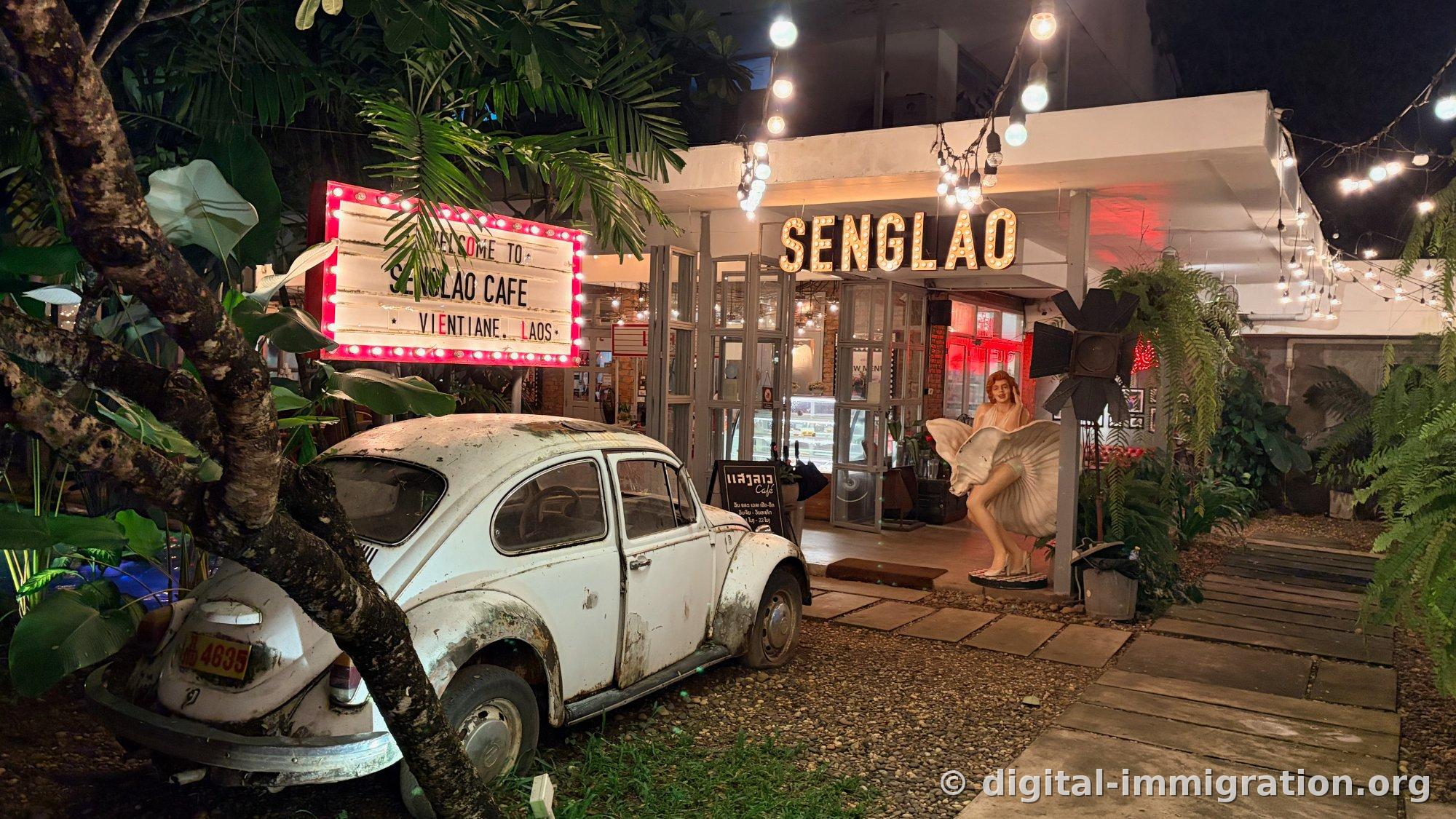 Vue nocturne de l'extérieur du café SENGLAO, l'enseigne au néon, l'enseigne en relief SENGLAO, la Coccinelle blanche dans la cour avant et la statue de Marilyn Monroe