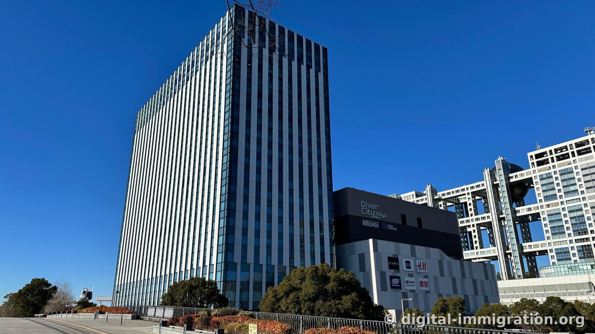 A glass curtain wall high-rise office building on the right with DiverCity Tokyo Plaza signage and logos for ZARA·GU·H&M·SKECHERS·ABC-MART, the Gundam's upper half small and nestled above trees on the lower left