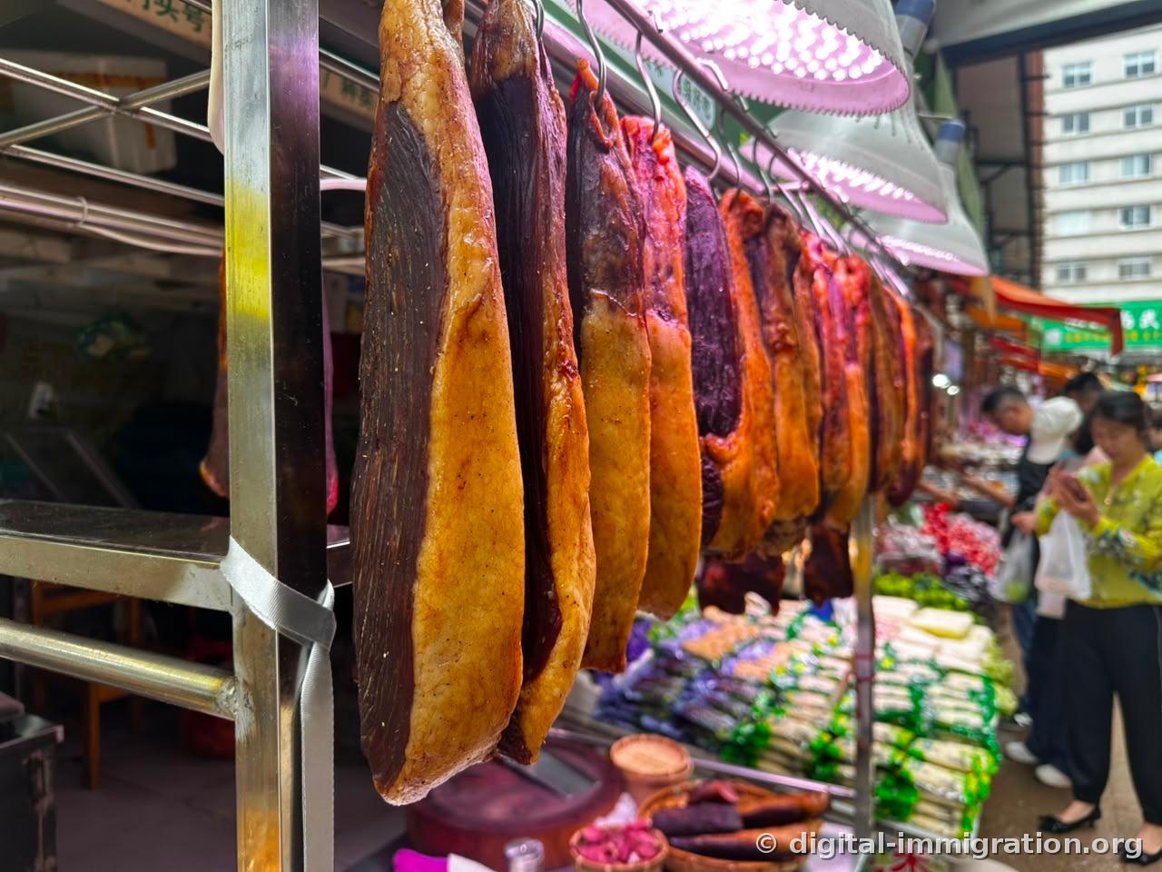Cured meat hanging in a line from hooks on stainless steel rods in a market alley, pink lighting from above, customers passing by