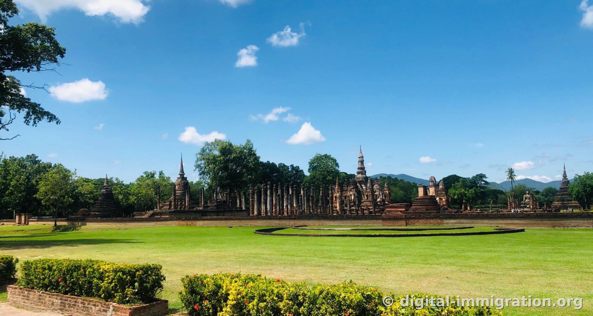 A wide grass field and columns and spires of Wat Mahathat stretching out beyond a yellow flower bed, with blue sky and white clouds
