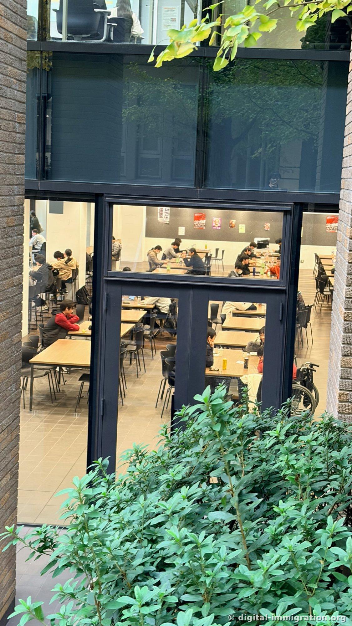 玻璃窗外学生食堂内部，长木桌前坐着进食的学生