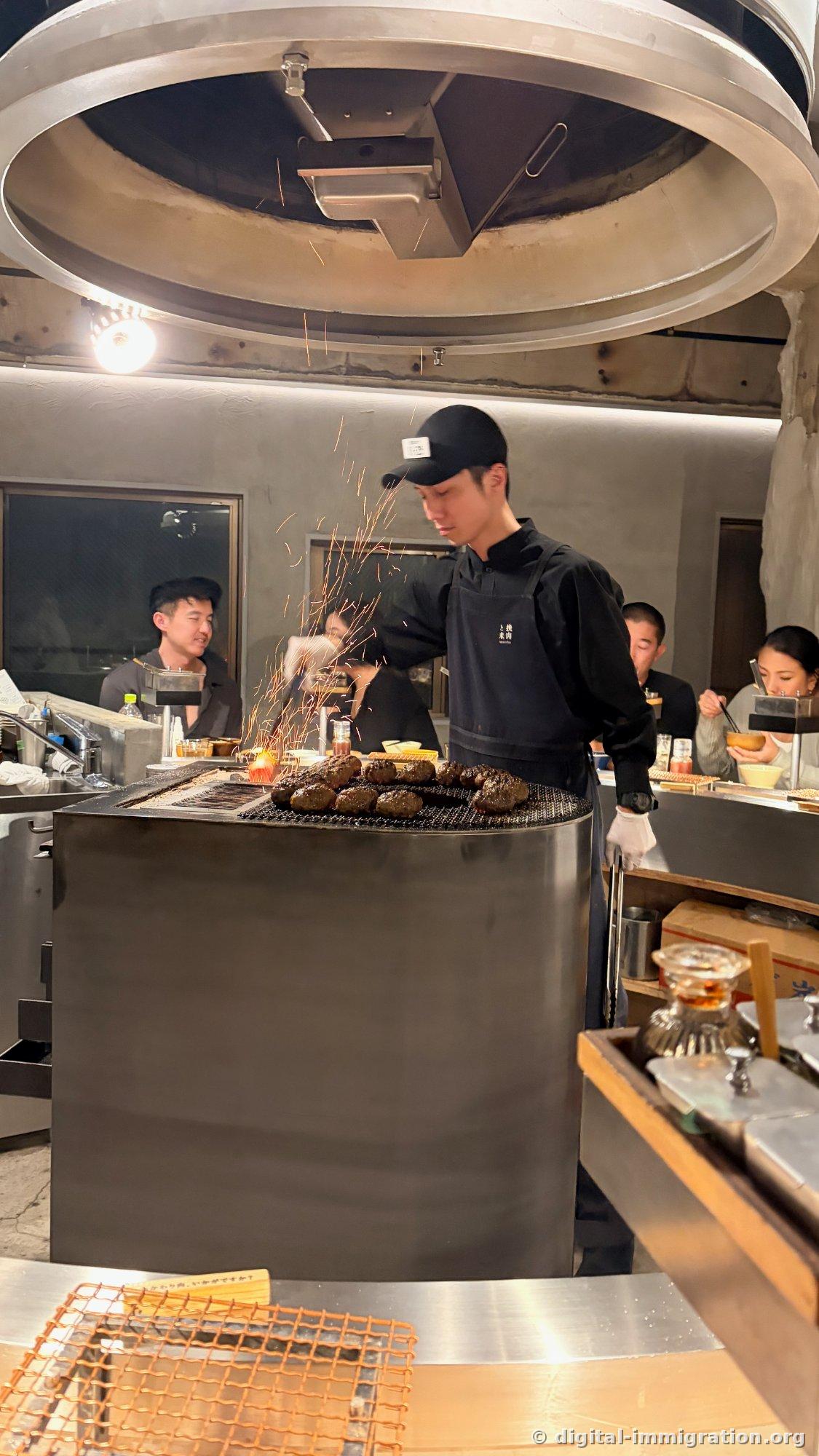 Un employé en tablier noir et chapeau grille plusieurs hamburgers sur le foyer de charbon central du comptoir, des étincelles jaillissent tandis que les clients regardent