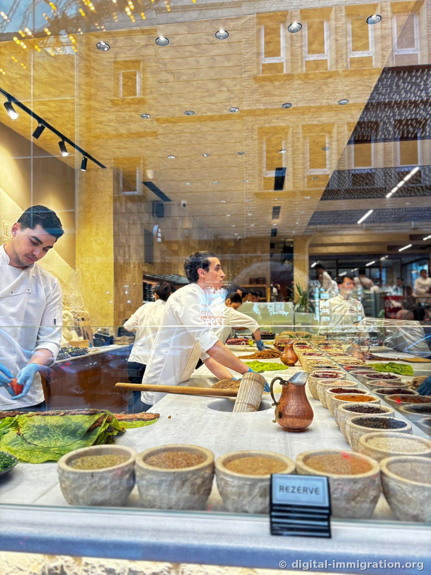 Through a glass window, two staff members in white uniforms and spices in stone bowls, a copper kettle and REZERVE sign on the dessert shop counter