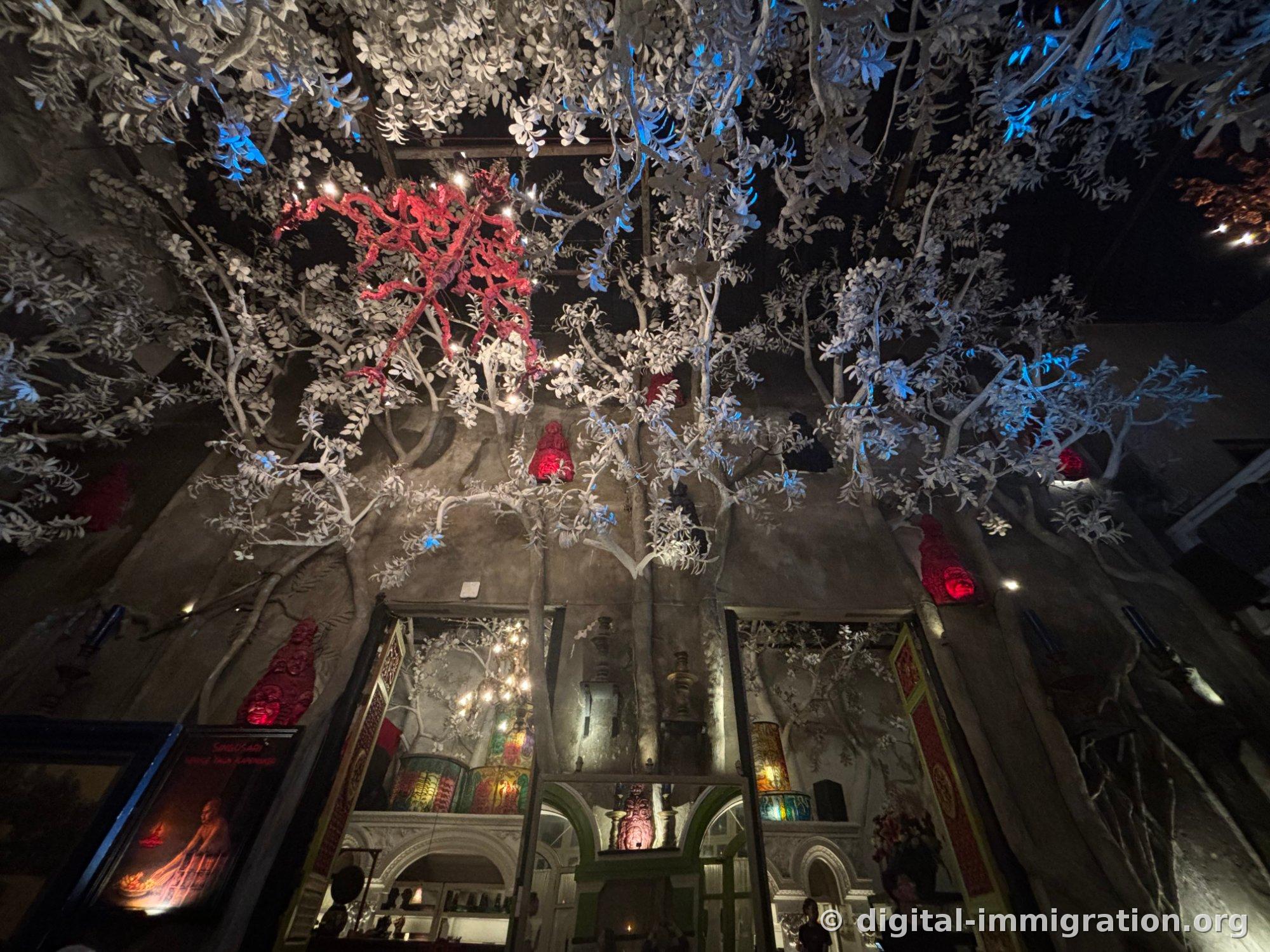 A massive tree sculpted in gray concrete covering walls and ceiling of a hall, with red coral-shaped chandelier and blue lighting