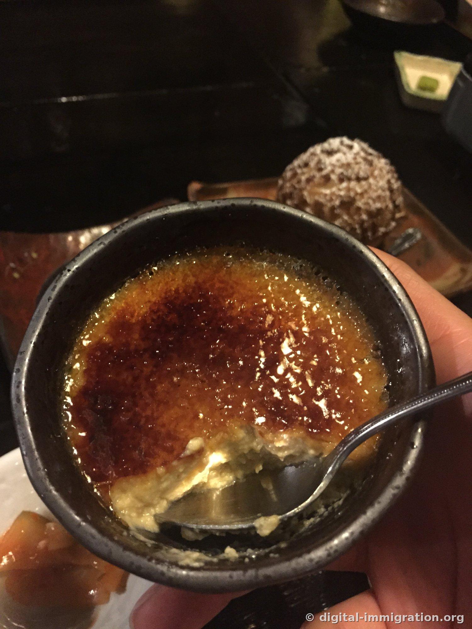 검은 도자기 그릇에 담긴 흑당푸딩, 표면이 캐러멜라이즈되어 갈색으로 그을려 있고 숟가락으로 파낸 자국, 뒤쪽에 슈크림 빵