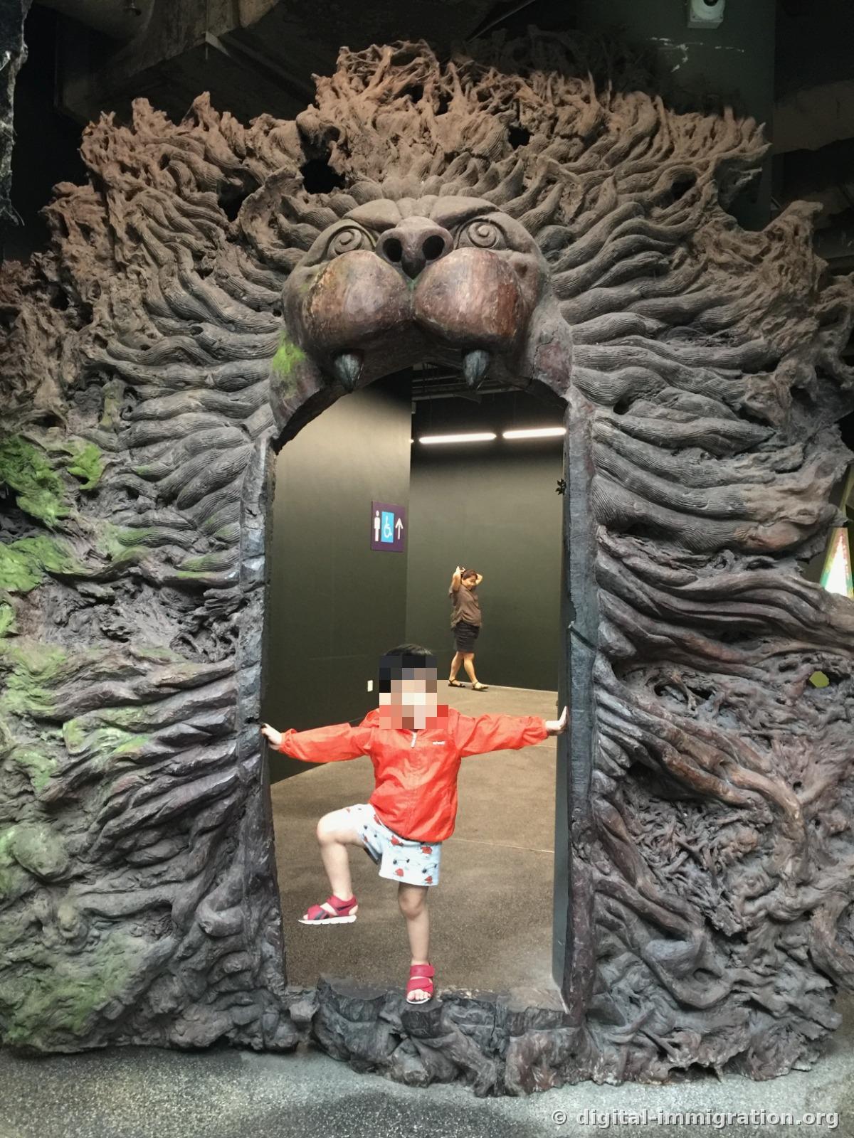 A passageway entrance carved in the shape of a beast's mouth with tree roots, with a child standing in front with arms spread, and a restroom sign visible inside