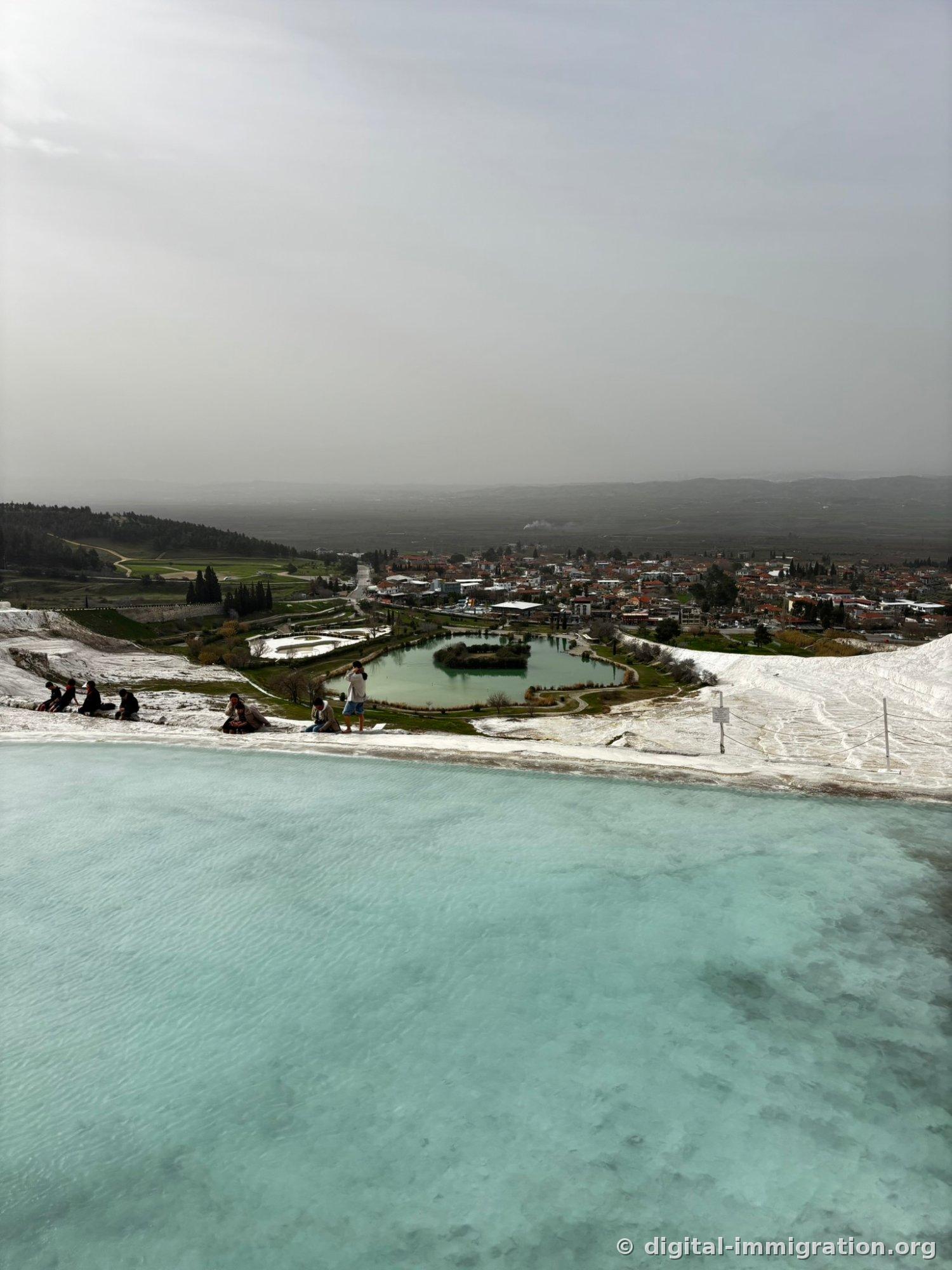Beyond mint-colored shallow hot spring water, people sit on the edge of the travertines, with a green lake and the orange-roofed village of Pamukkale visible below