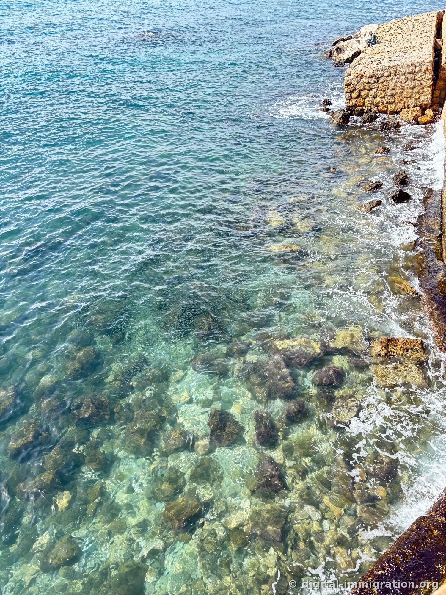 Crystal-clear turquoise shallow water with visible bottom rocks, person sitting alone on stone wall above right