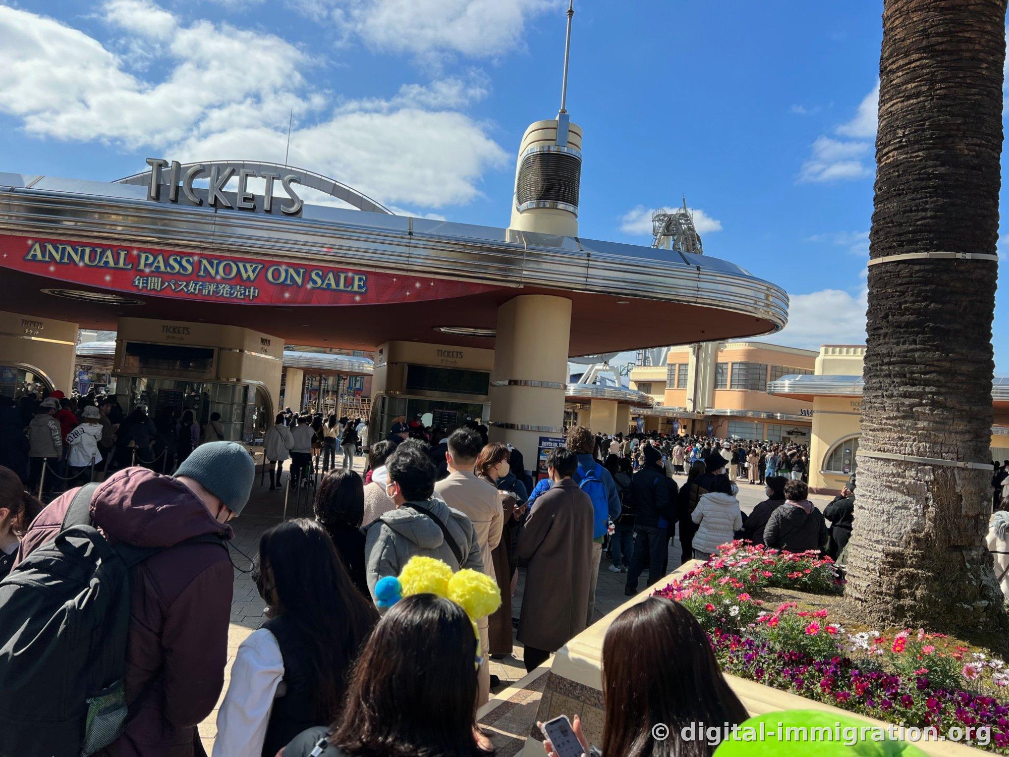 Long lines of visitors queue in front of the Universal Studios Japan TICKETS booth with a red annual pass promotion banner hanging above