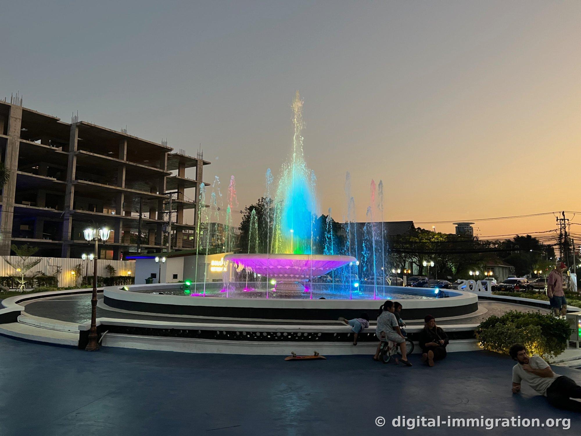 Ciel orange après le coucher du soleil avec la fontaine Nam Phou illuminée en bleu, vert et rose, des enfants assis au bord, des skateboards sur le sol de la place
