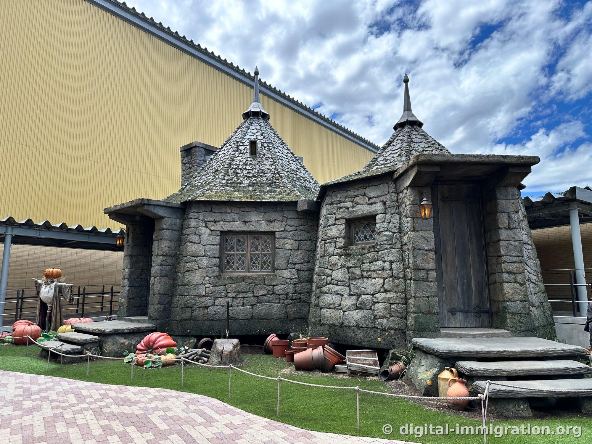 An outdoor stone hut with a conical roof, surrounded by large orange pumpkins and a scarecrow