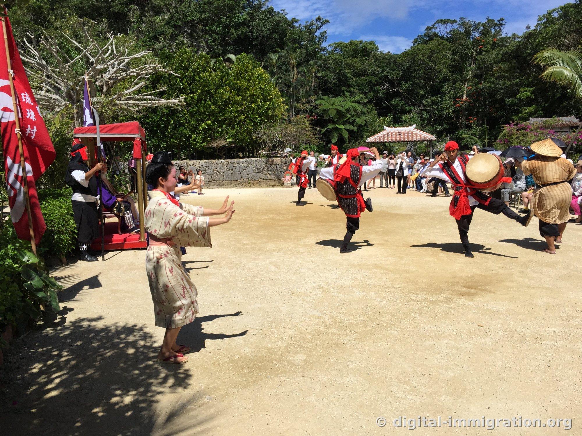 Un tambourinaire Eisa portant un foulard rouge lève haut ses jambes en sautant, tandis qu'une danseuse vêtue d'un kimono fait des gestes de main devant