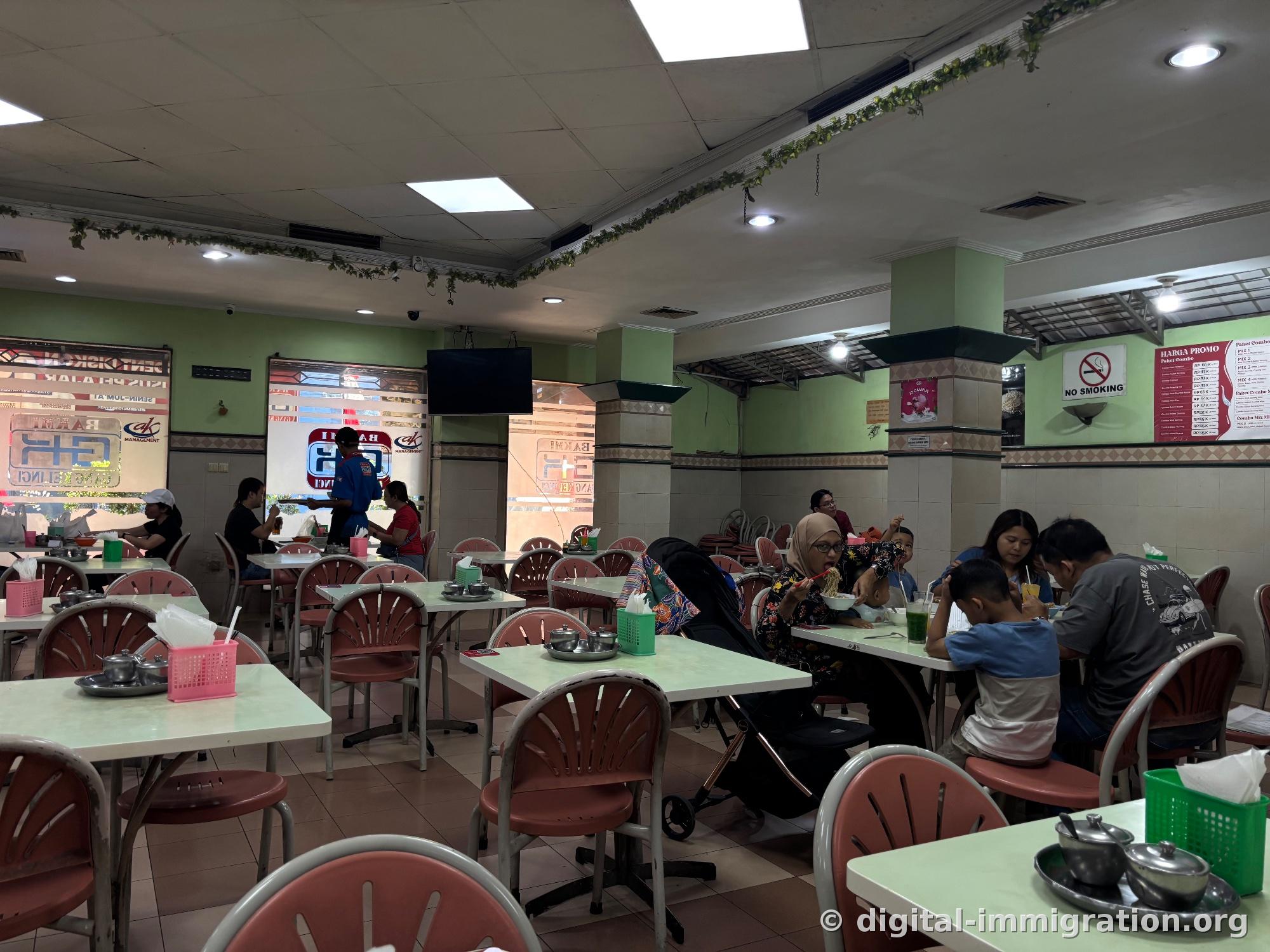 Vaste salle du restaurant avec chaises roses et tables blanches, familles qui mangent des nouilles au fond, tableau de prix HARGA PROMO et panneau NO SMOKING sur les murs
