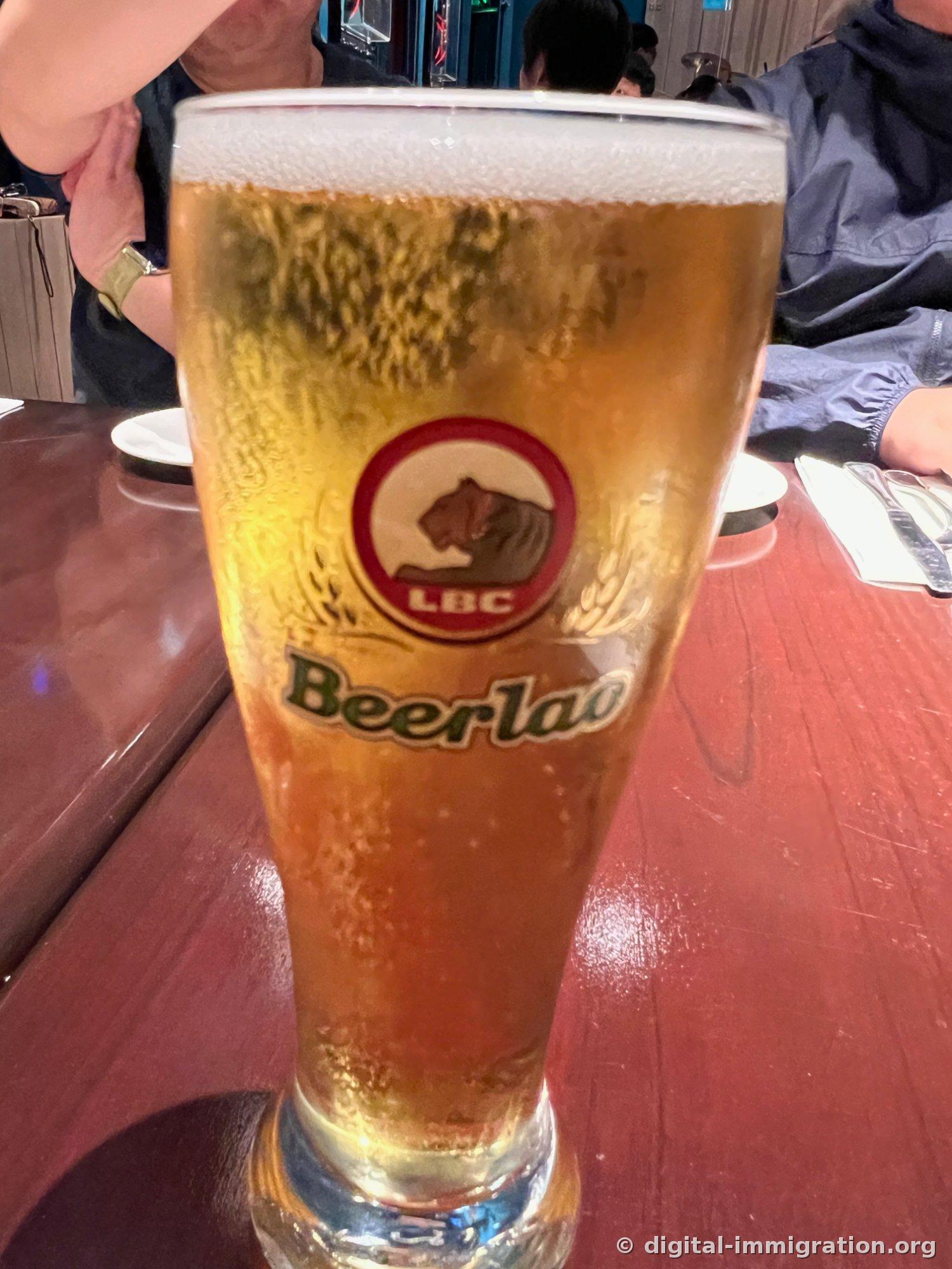 붉은 원 안 호랑이 머리 LBC 로고와 Beerlao 글자가 새겨진 잔에 거품 올라온 생맥주가 가득 담겨 나무 테이블 위에 놓여 있다