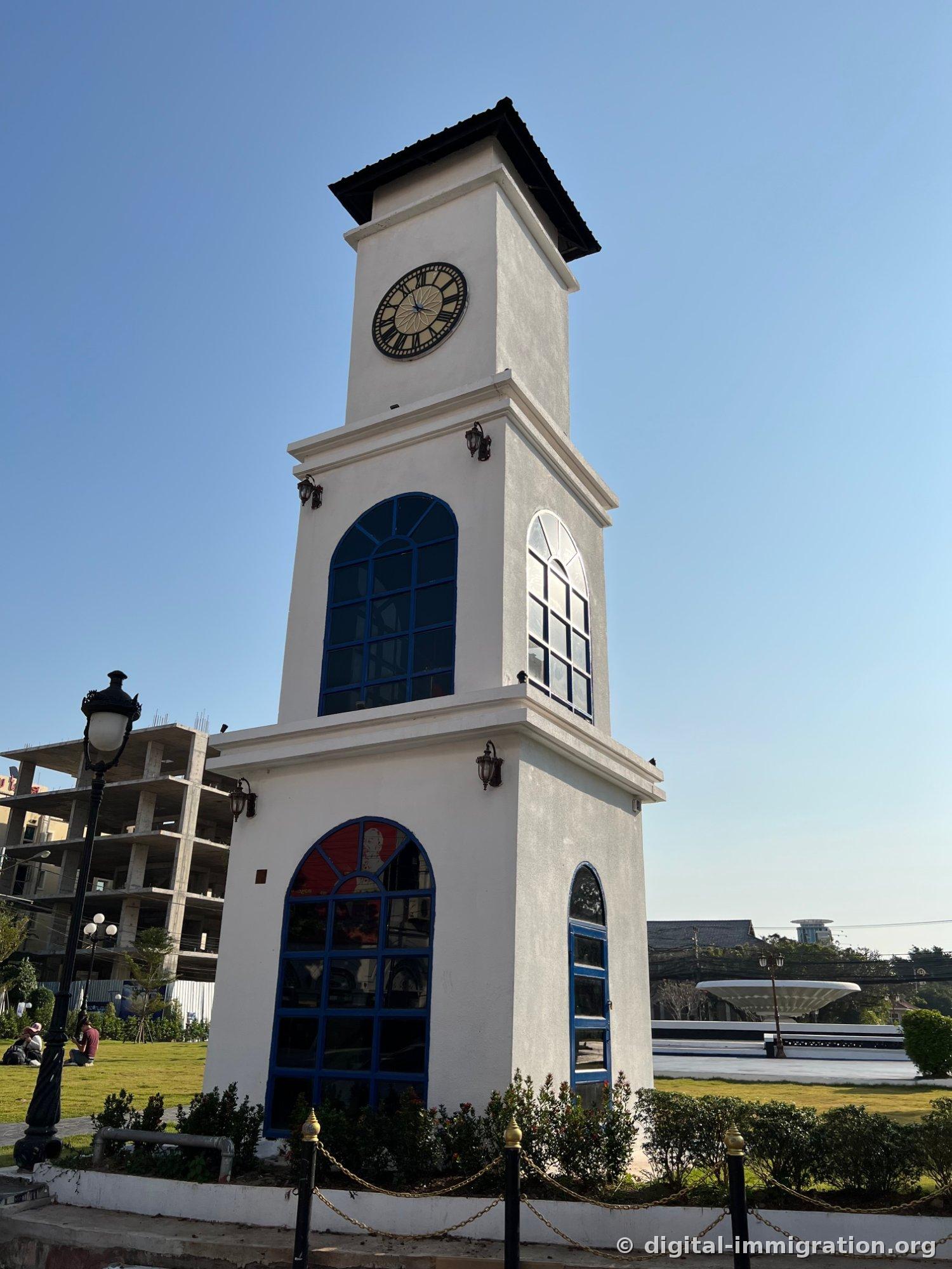 Une tour horloge blanche avec des arcs bleus aux fenêtres et un cadran romain, avec un immeuble de béton inachevé aux armatures visibles en arrière-plan