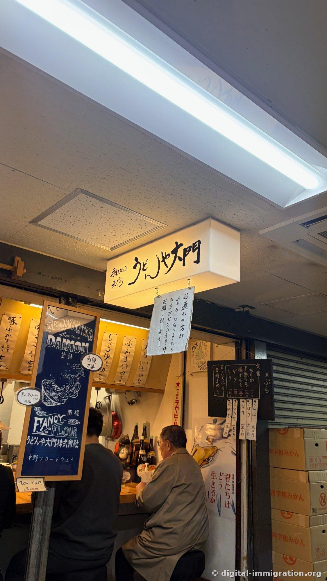 『手打うどん 天ぷら うどんや大門』の照明看板とその下に『一行がそろったら列に並んでください』という手書きの案内文が掛かる店入口