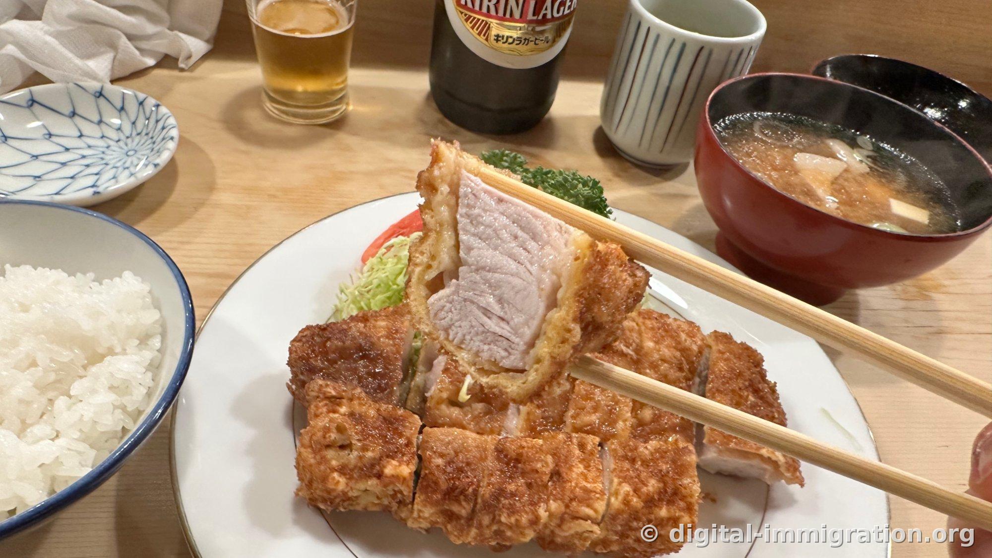 A cross-section of roast cutlet held up with chopsticks, showing the thin breading surrounding pale pink pork