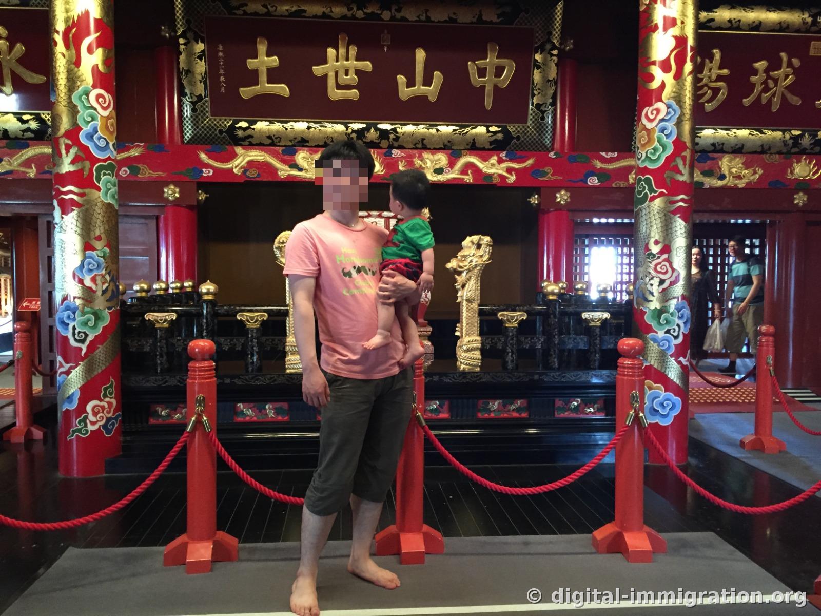 Holding a child barefoot in front of the royal throne of Shuri Castle Seiden with the plaque reading '中山世土' and golden dragon pillars