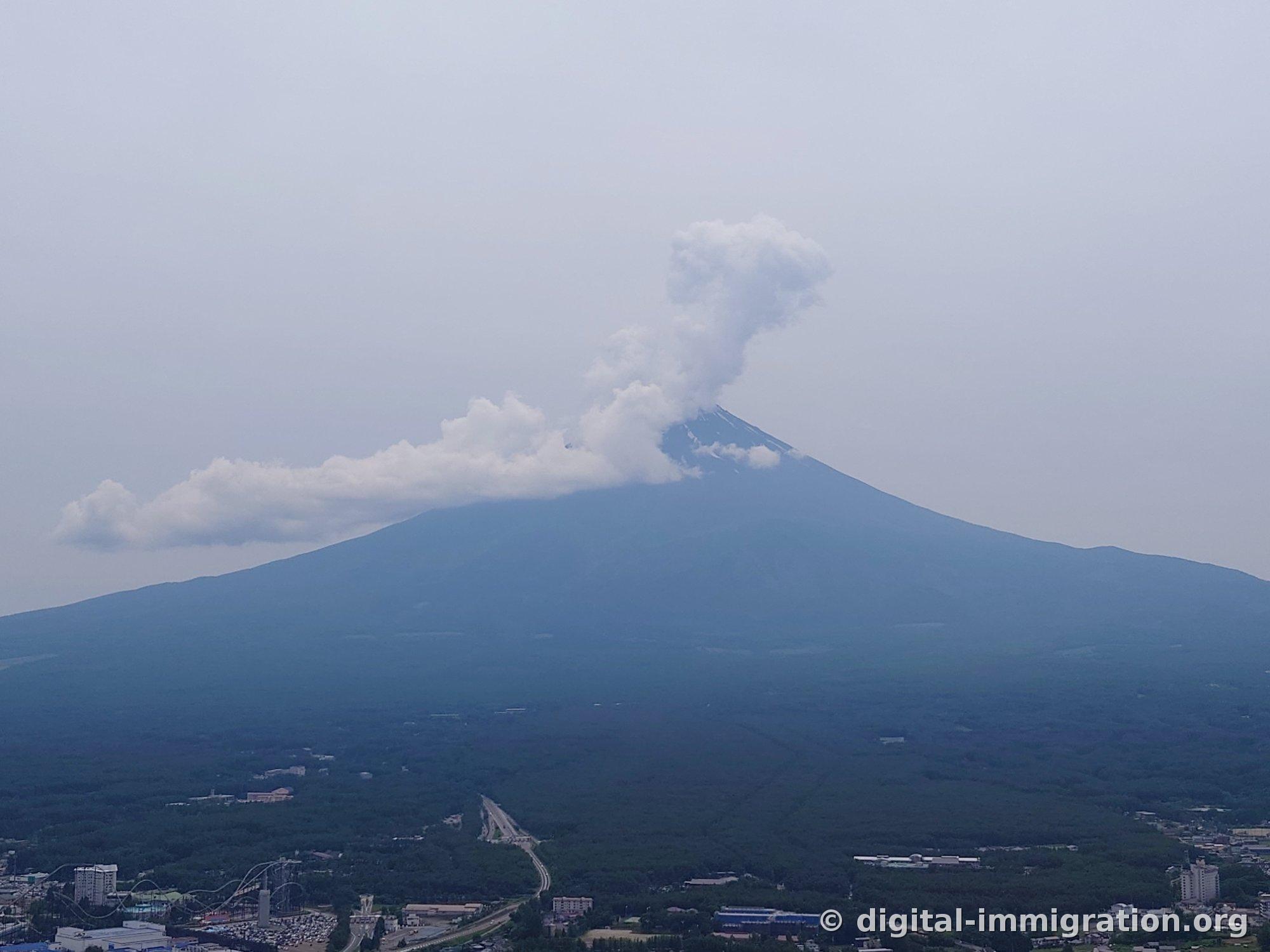 A close-up of Mount Fuji's summit with white clouds billowing up, and roller coaster rails visible at the base