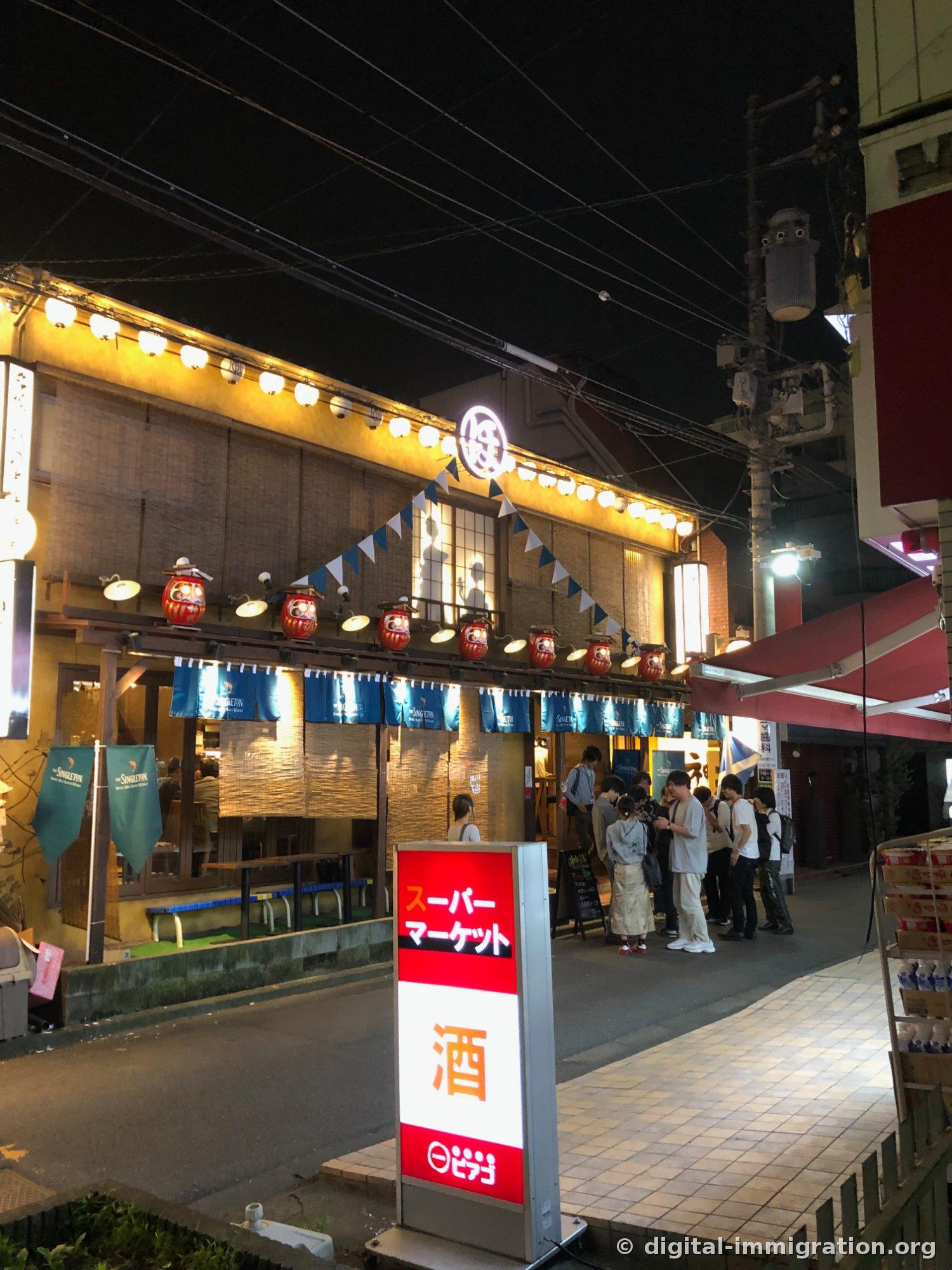 达摩灯笼排成一列的居酒屋外观，前面竖着红色超市招牌