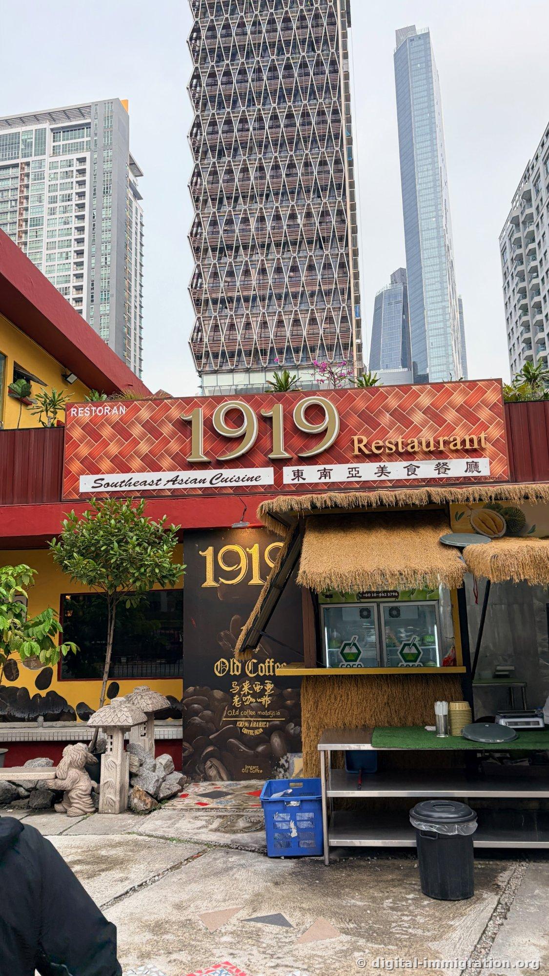 The restaurant's front entrance, featuring a red signboard with '1919 Restaurant' in gold lettering, with Southeast Asian Cuisine and Chinese characters 東南亞美食餐廳 below, and a straw-roofed coconut stand with refrigerator beside it