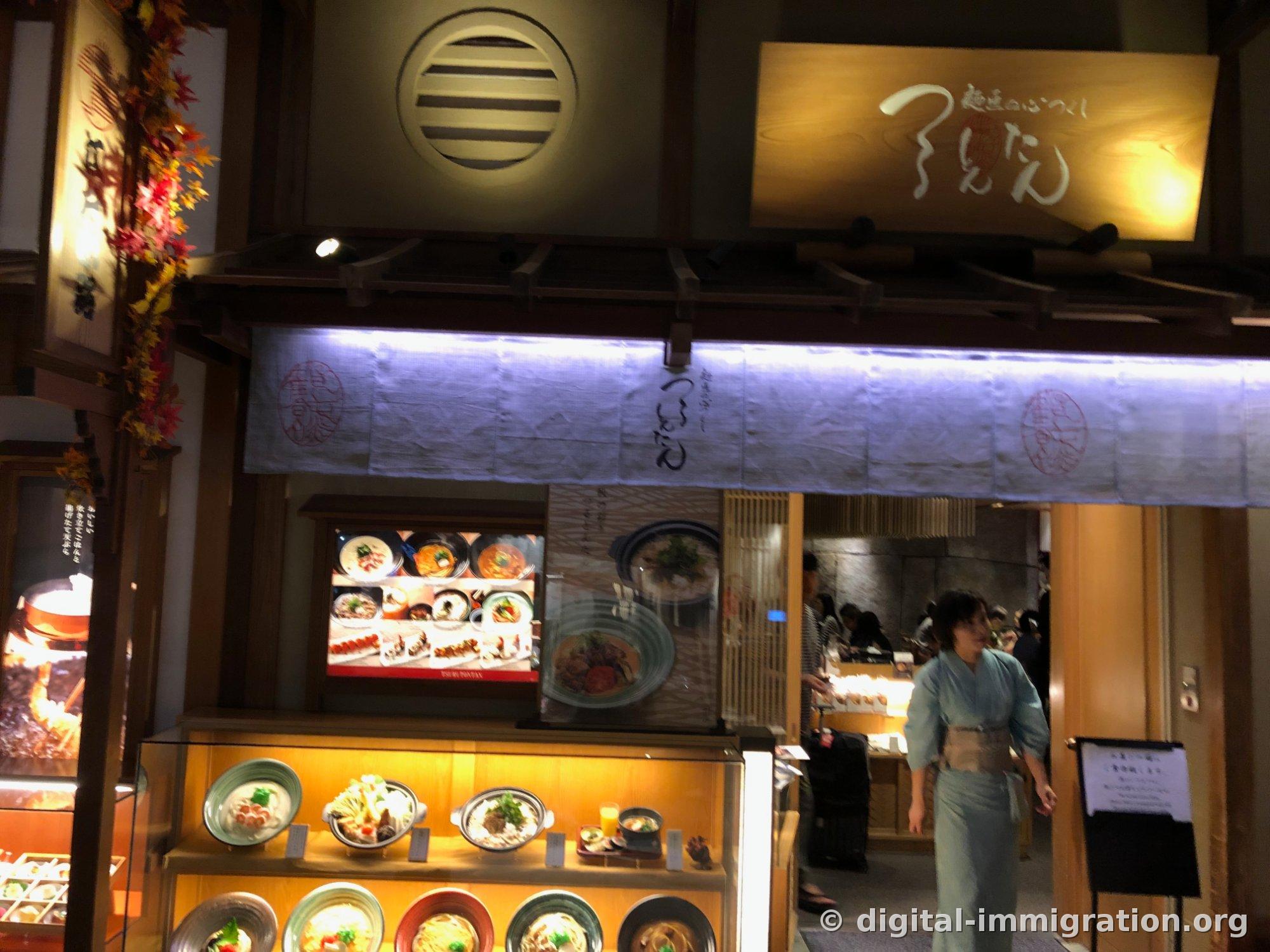 Entrance of Tsurutontan Haneda Airport branch with wooden eaves and white noren curtain, with a staff member in light blue kimono passing on the right