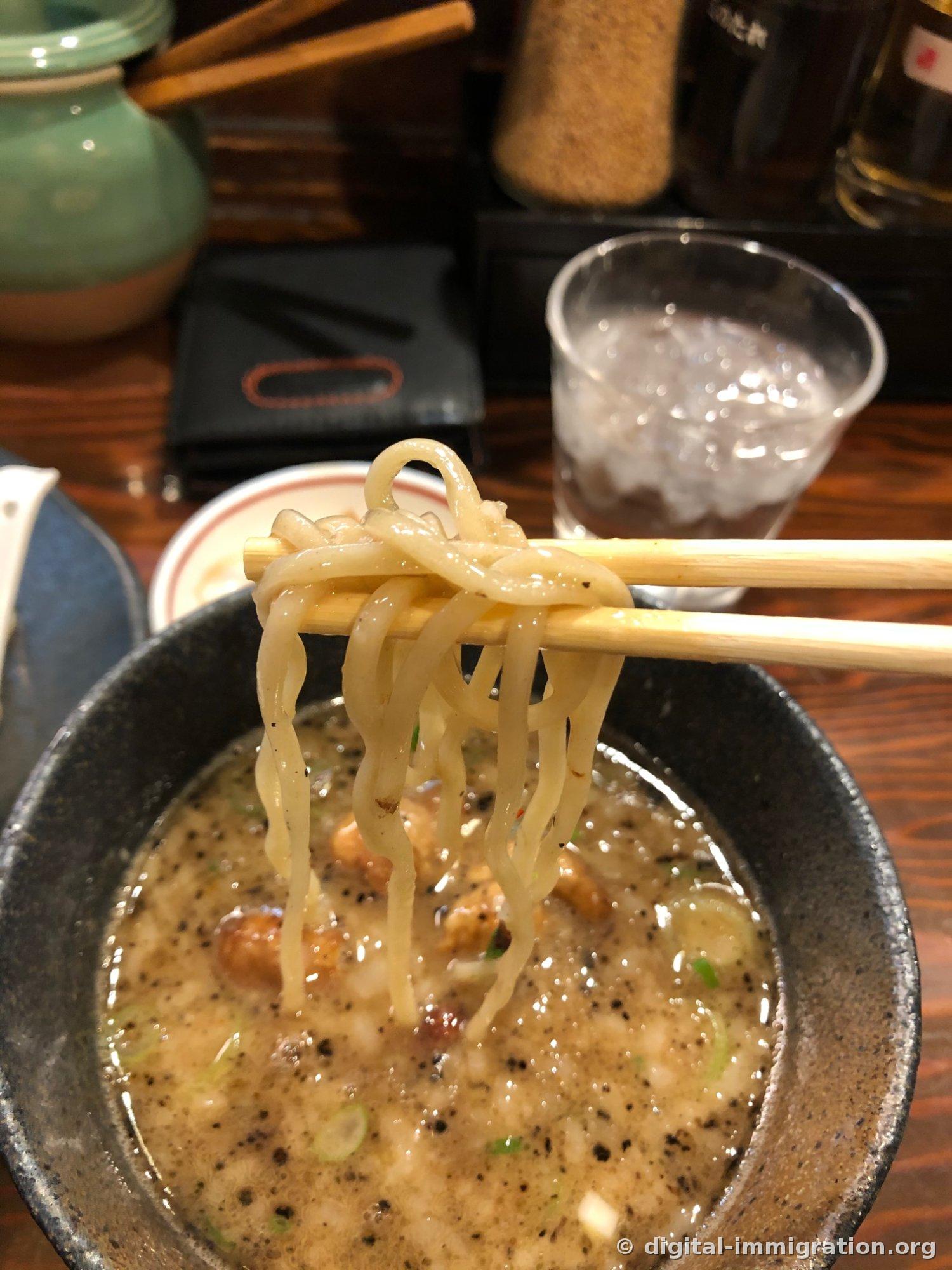 Les nouilles soulevées du bol du bouillon avec des baguettes. Le bouillon épais et les particules noires de shoyu brûlée adhèrent aux nouilles, avec le motsu et les oignons verts visibles dans le bol