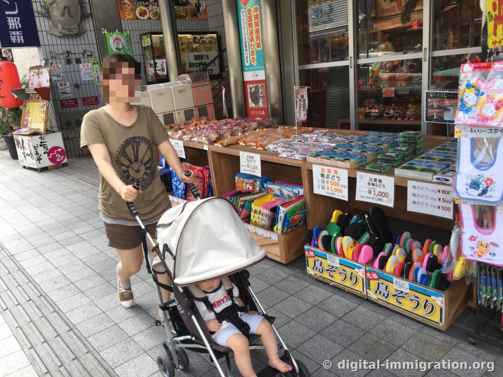 Sur l'étalage devant un magasin de Kokusai-dori, des boîtes de raisin de mer et des pantoufles Shimajori multicolores, une personne poussant une poussette qui passe