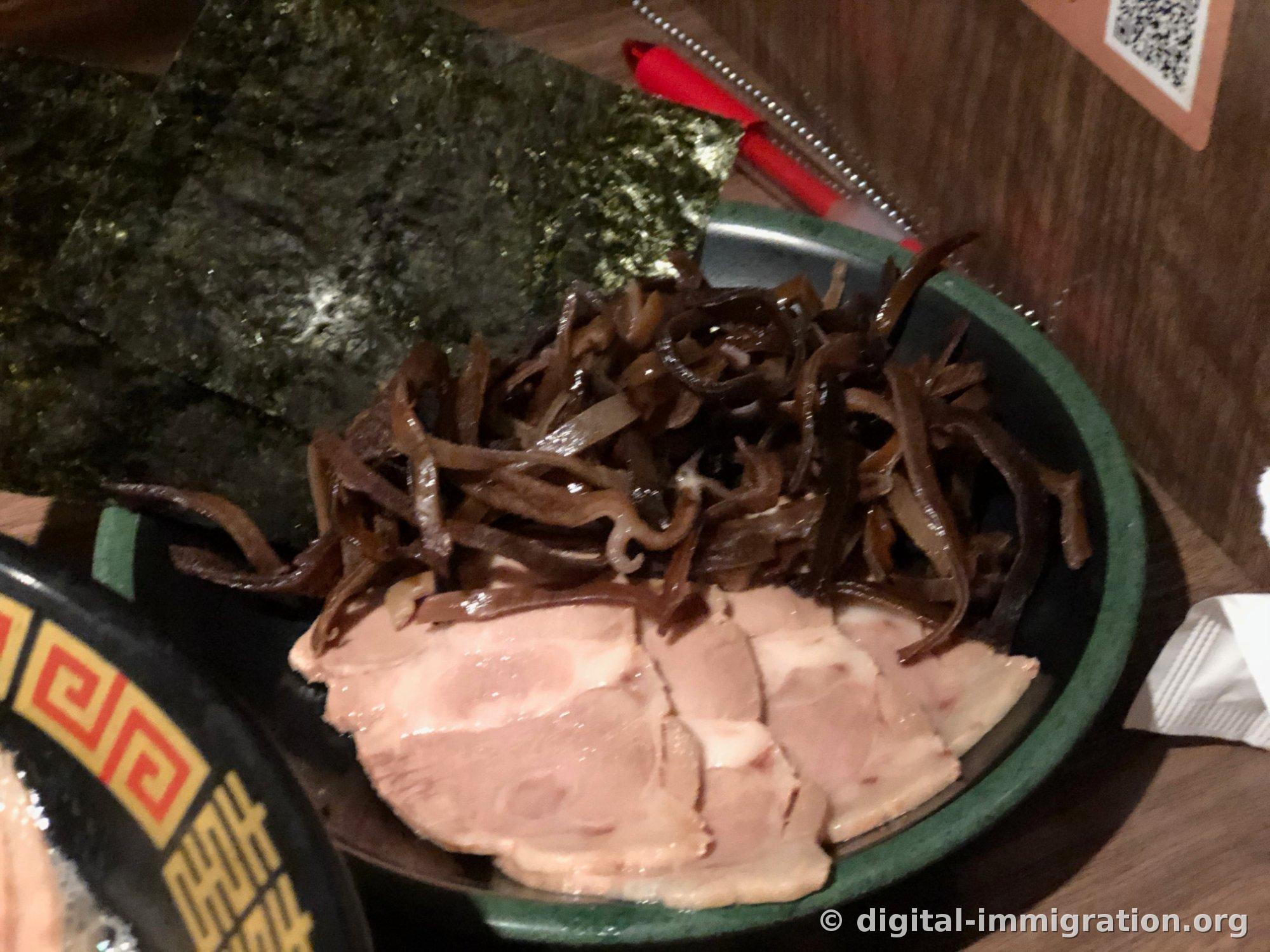 Une fine tranche de char siu soulevée du bouillon avec des baguettes, avec un œuf fendu en deux visible en bas du bol
