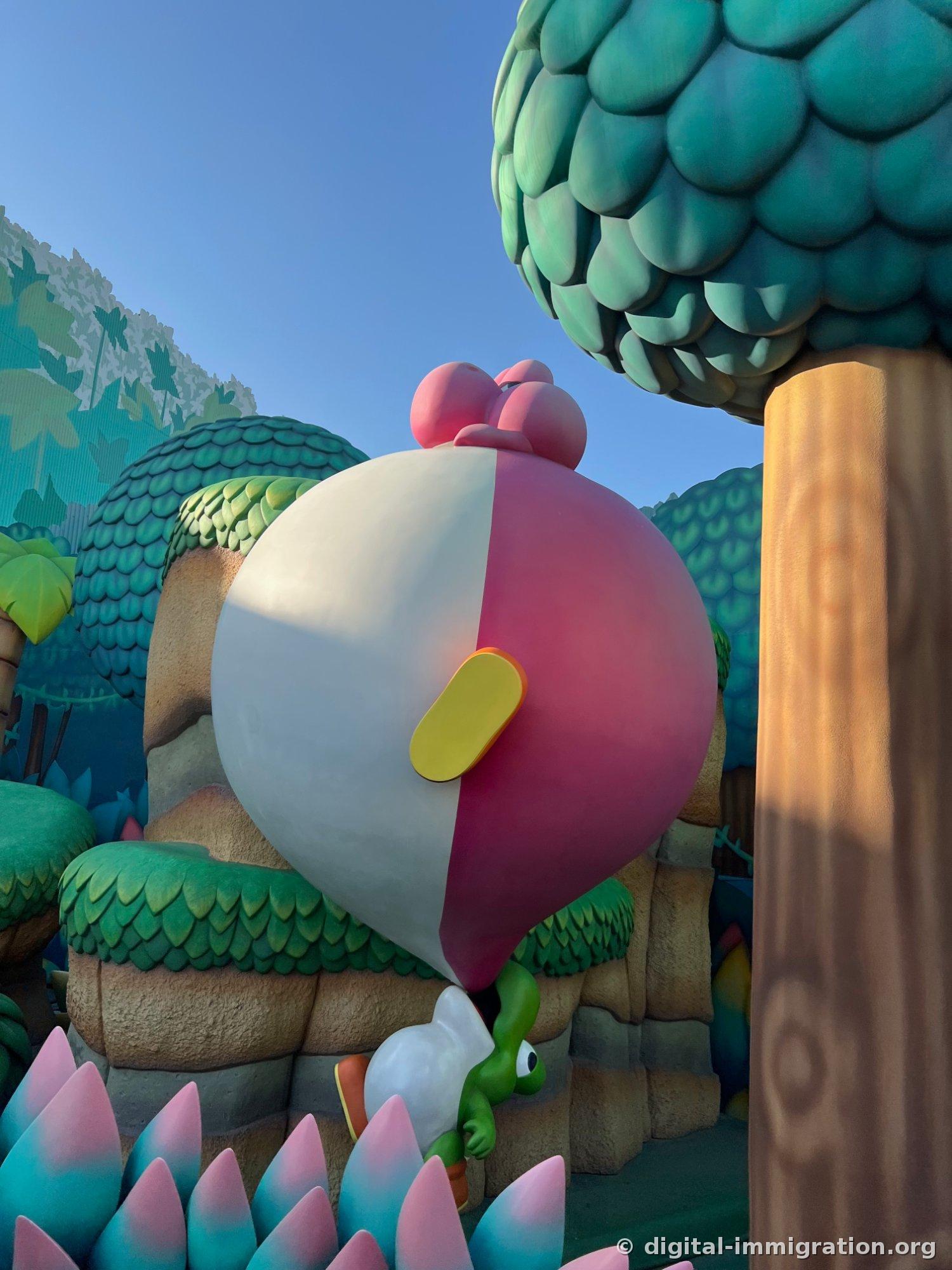 A pink Yoshi peeks out from behind a giant egg sculpture divided in pink and white, with a green Yoshi and pastel spiky plants below