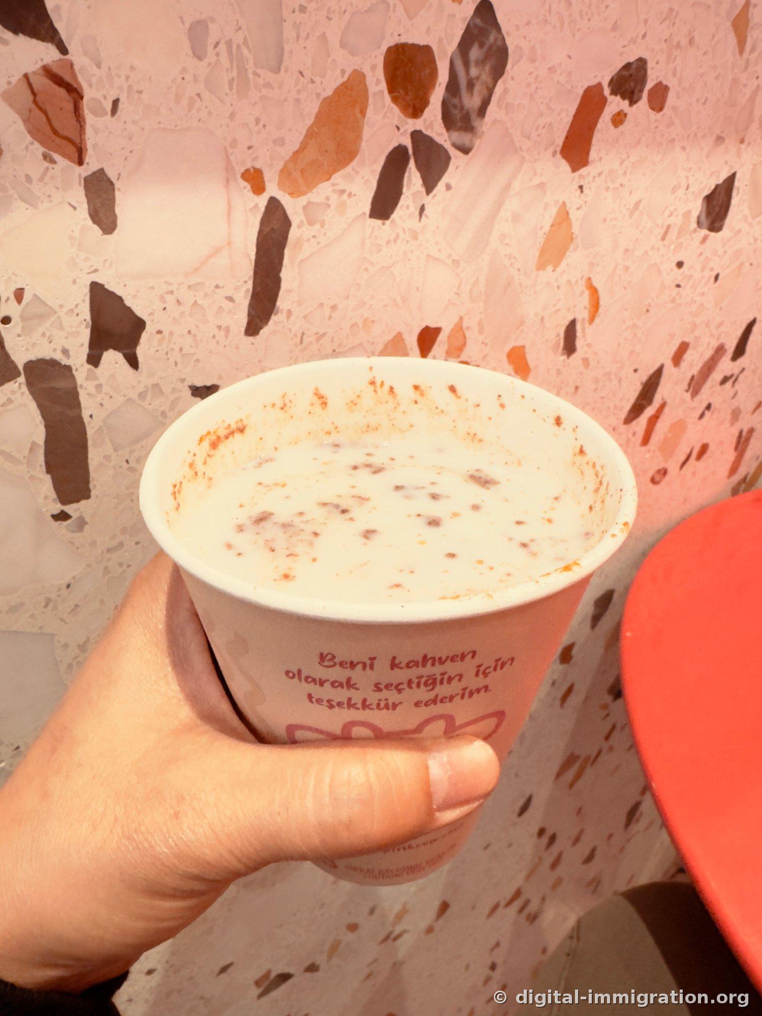 Gobelet en papier tenu à la main contenant du salep blanc saupoudré de cannelle. Sur le gobelet, le texte turc 'Beni kahven olarak seçtiğin için teşekkür ederim', fond d'un mur en terrazzo rose