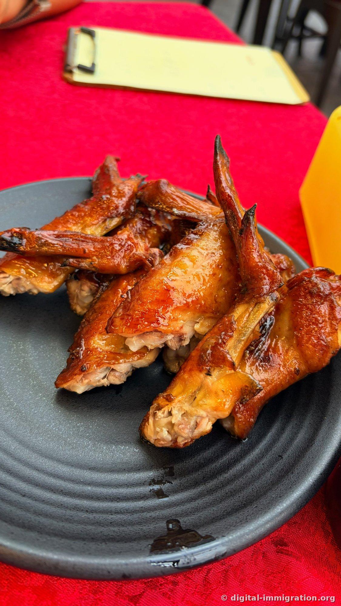 つやのある茶色に焼き上がった鶏の手羽がいくつか黒いお皿に盛られ、後ろに黄色いメニュー板と赤いテーブルクロスが見える光景