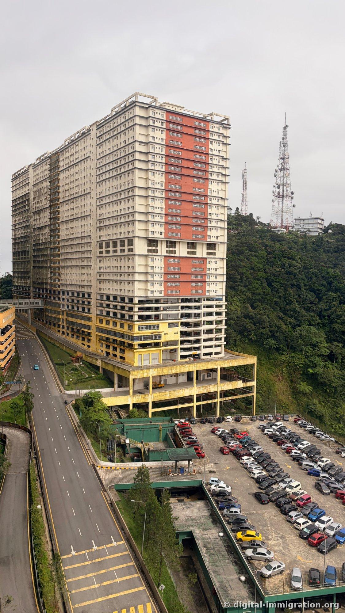 Large white and red residential building standing on mountain ridge, communications tower on adjacent hill, densely packed cars in open-air parking lot below