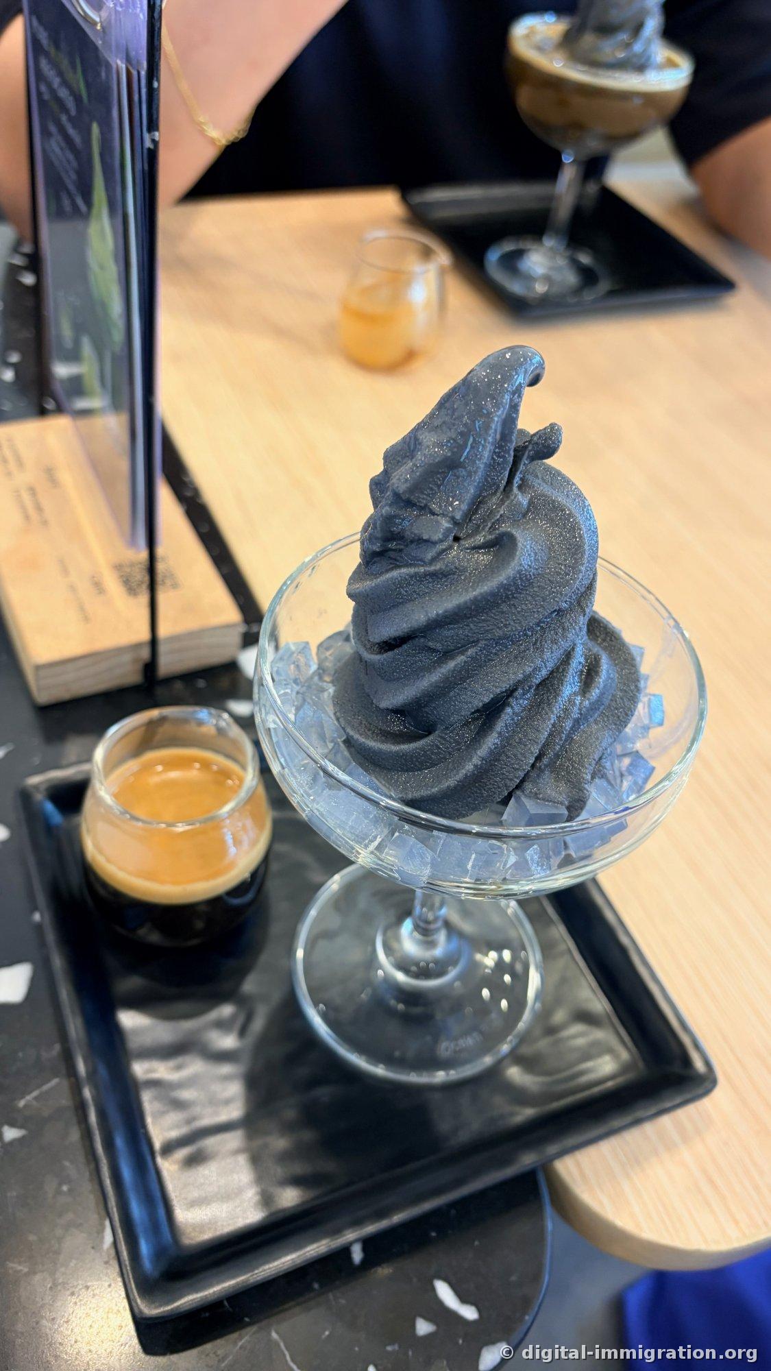 Jet-black soft serve ice cream with transparent jelly cubes in a glass on a black tray, with a small glass of espresso beside it, and the same dish visible on a nearby table in the background