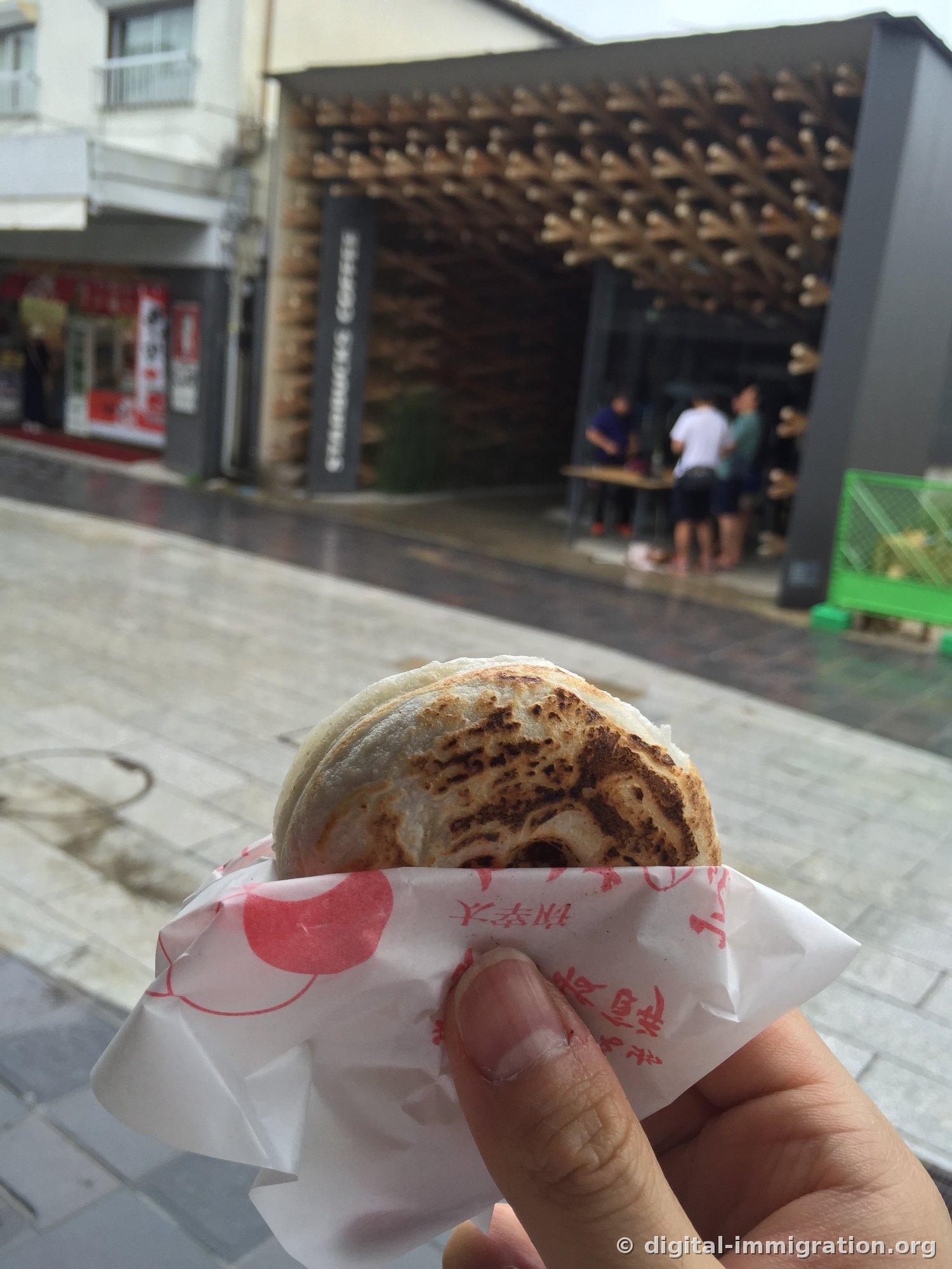Freshly grilled umegaemochi wrapped in plum-patterned paper held in hand, with a wooden lattice-facade Starbucks in the background