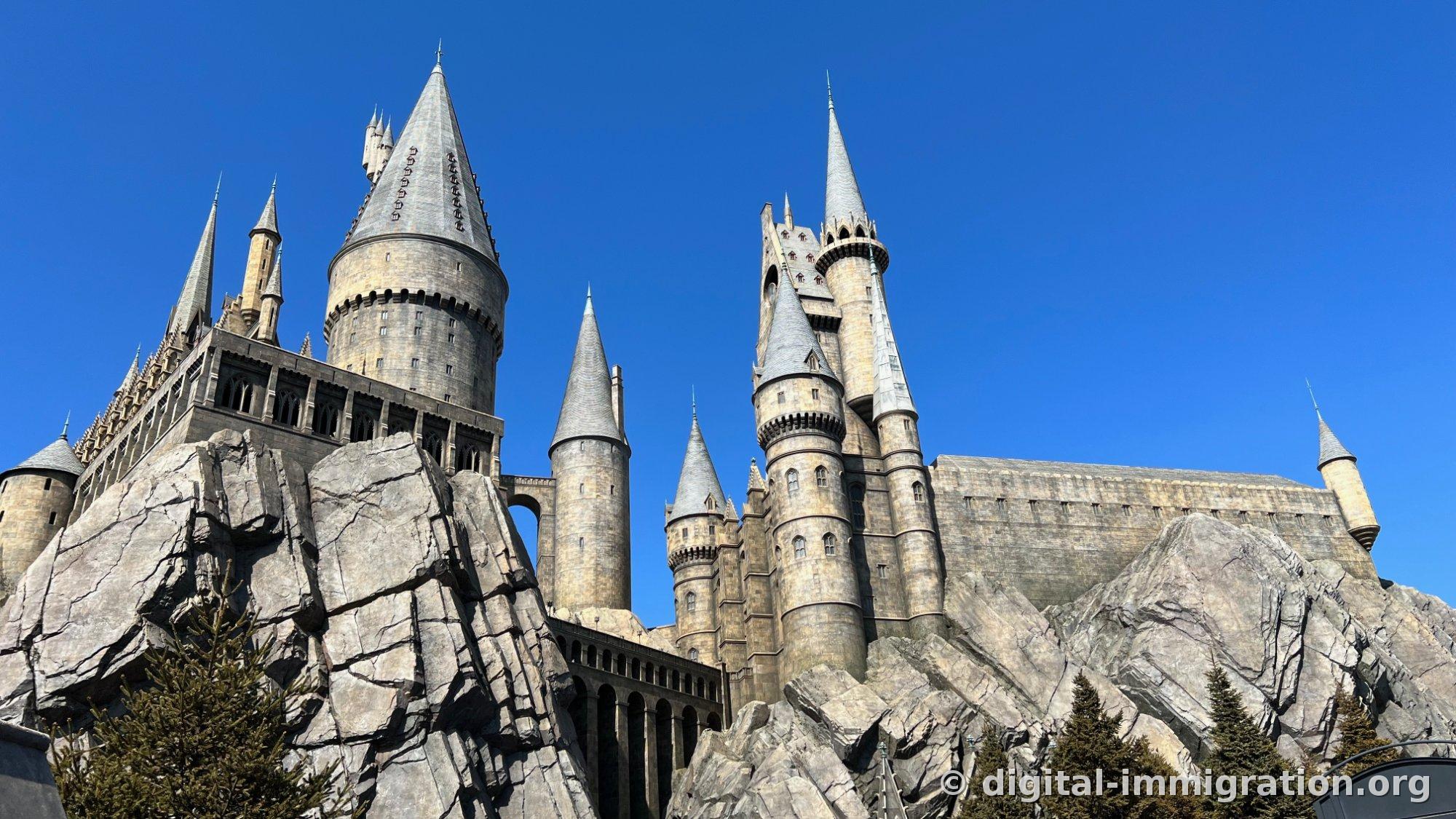 Hogwarts Castle seen from below atop a rocky mountain, with sharp gray spires rising against a clear blue sky