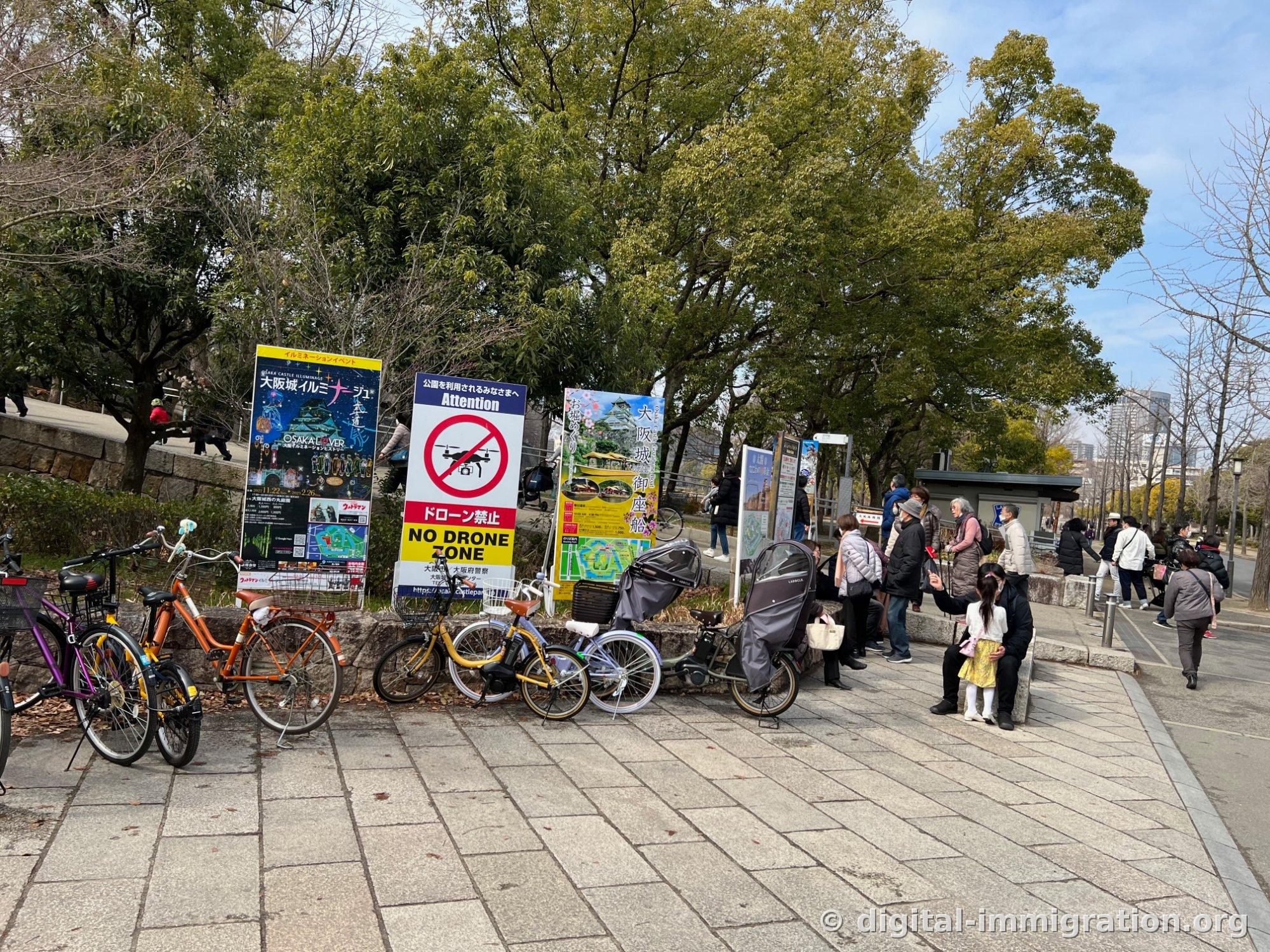 Vélos alignés sur le trottoir à l'entrée du parc du château d'Osaka, affiche d'illuminations, panneau d'interdiction de drones et affiche publicitaire pour la croisière Gozabune du château d'Osaka