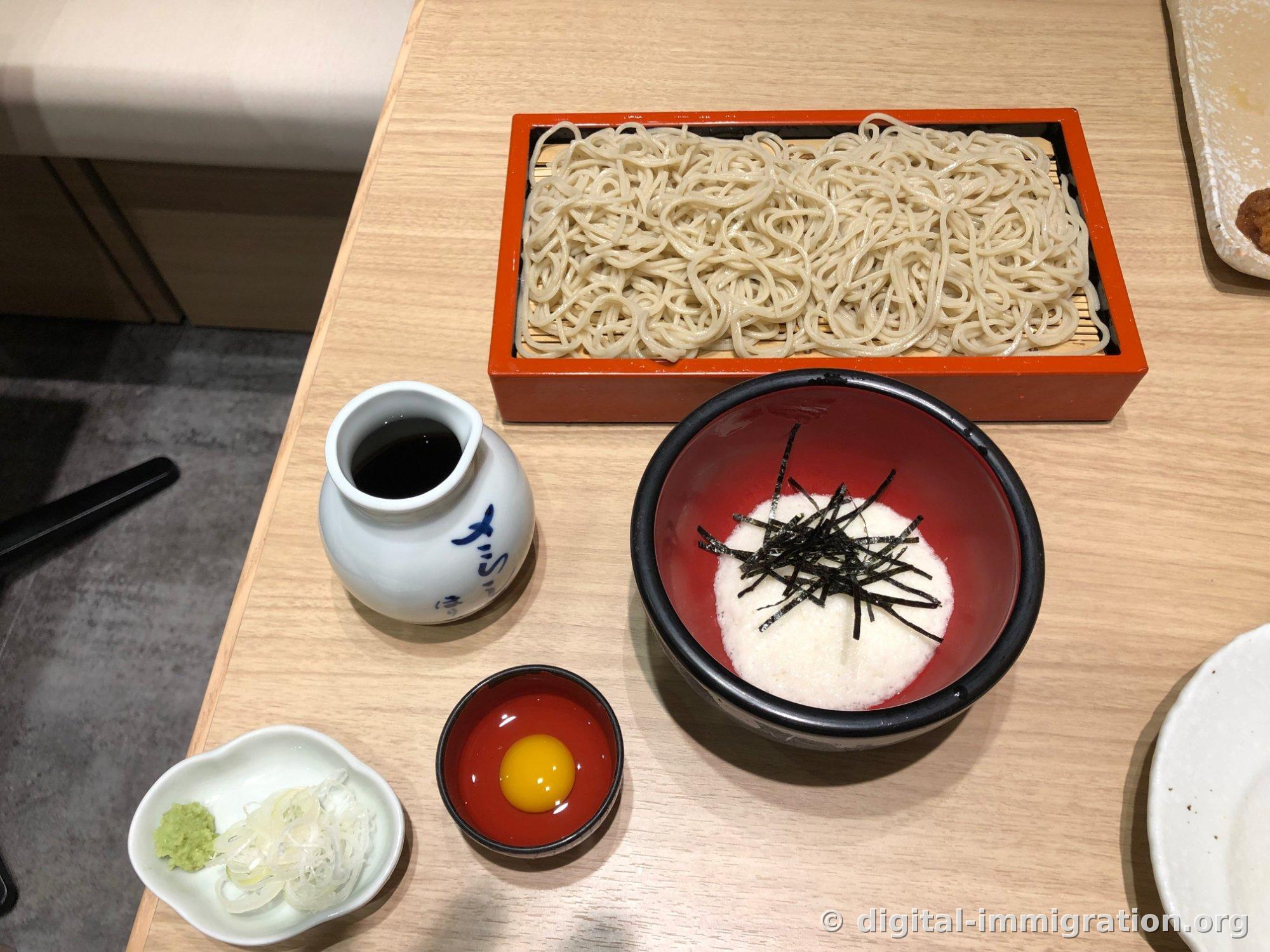 Soba in a crimson seiro, a white porcelain tsuyu pitcher, a dark tororo bowl with nori on top, a red dish with raw egg yolk, and wasabi and white scallions arranged on a wooden table