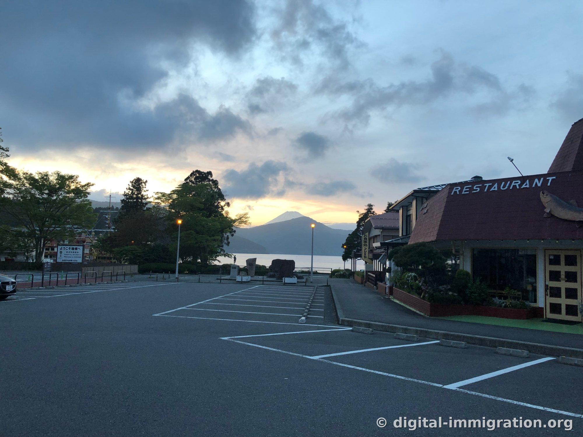 空荡荡的芦之湖停车场，停车场上方可见落日和富士山，右侧有红顶的RESTAURANT建筑和鱼形雕塑，左侧有'ようこそ箱根へ Welcome to HAKONE'招牌，傍晚景观