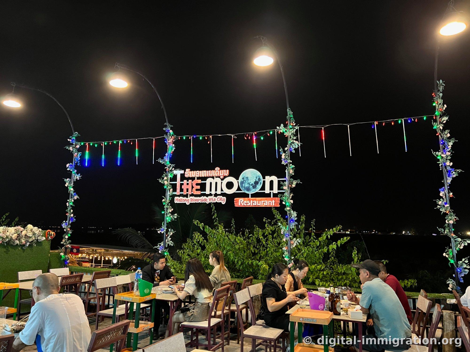 写着'THE MOON Restaurant, Mekong Riverside, Vte City'的月亮图案招牌，绿色和蓝色的串灯挂在露天餐桌区域上方