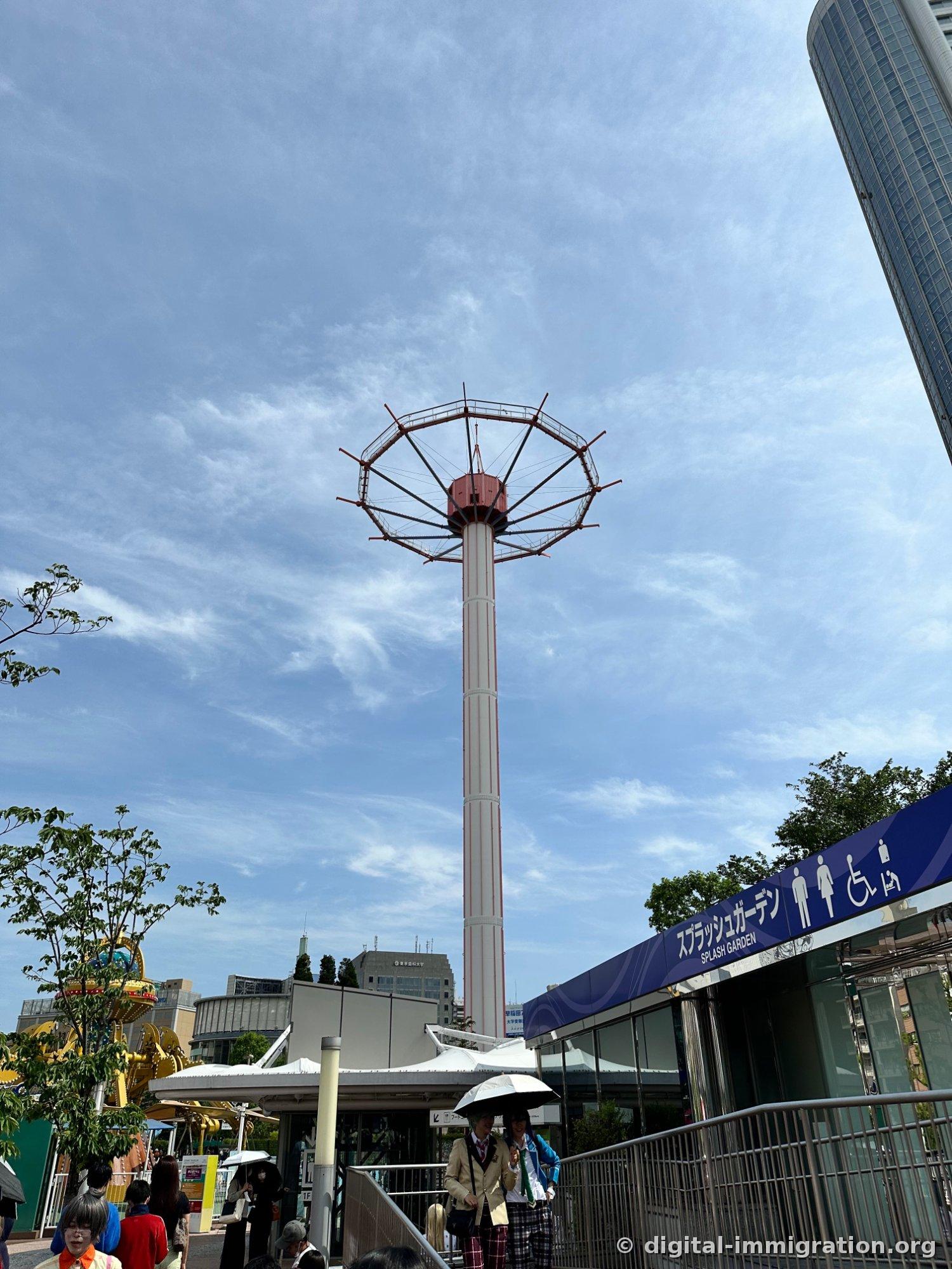Devant un ciel bleu, la colonne blanche d'une attraction de chute se dresse et sa partie supérieure circulaire rouge, avec un panneau bleu marine portant 'Splash Garden SPLASH GARDEN' en bas et des gens tenant des parasols