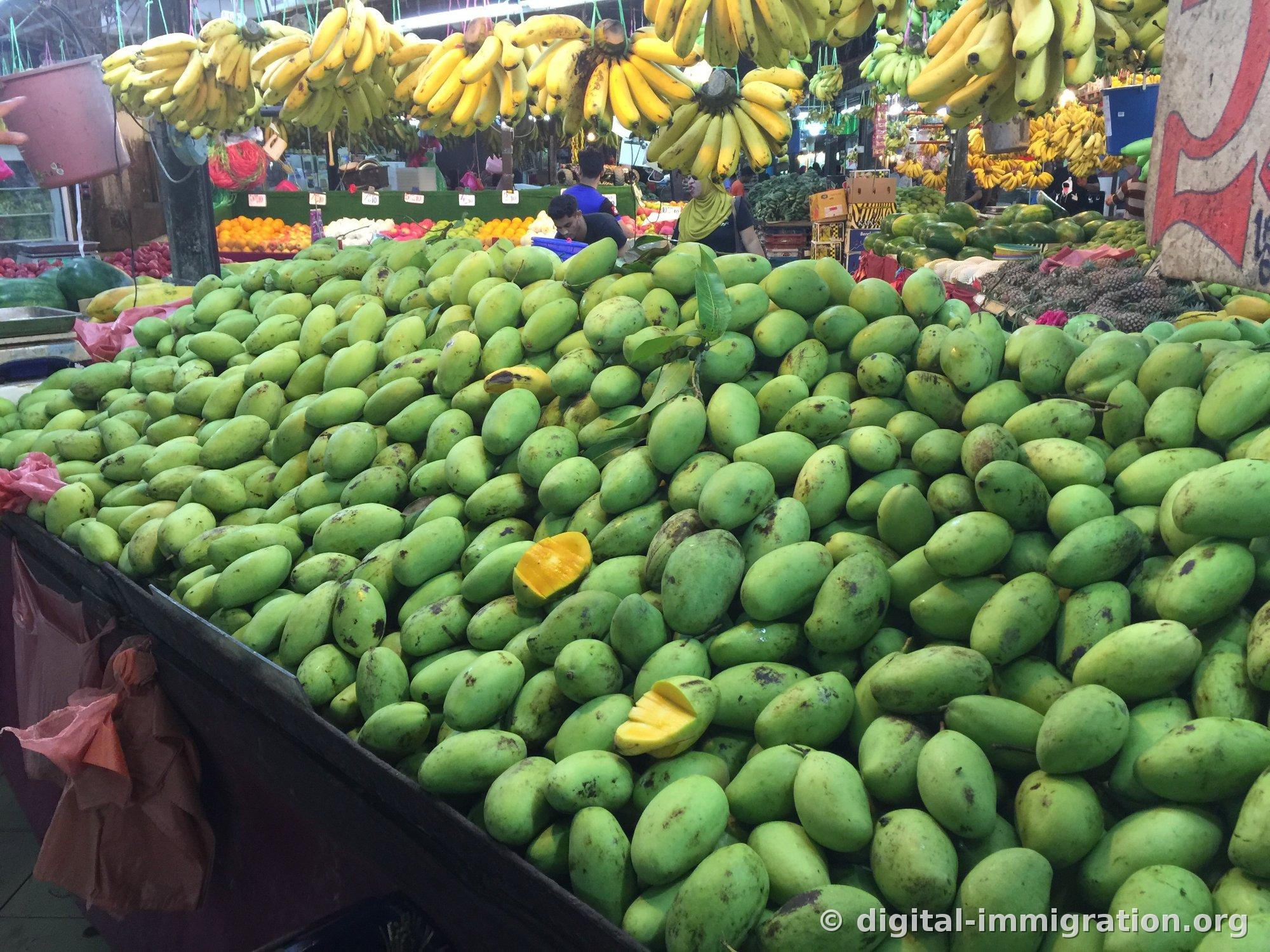 Vue générale d'un marché intérieur avec des mangues vertes empilées comme une montagne sur les étals du marché et des grappes de bananes jaunes suspendues au-dessus, avec des étals de pastèques et d'oranges visibles en arrière-plan