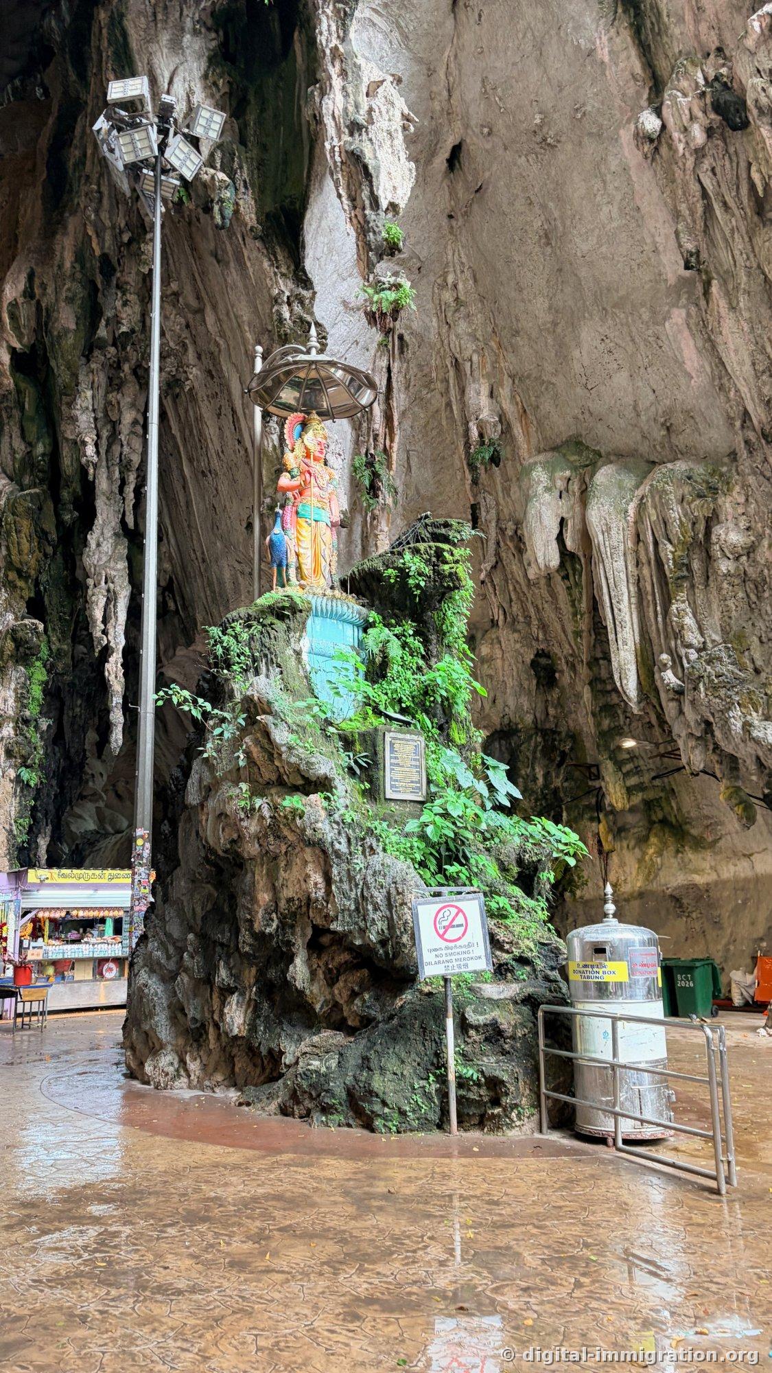洞窟の中の岩の上に建つムルガンの神像と孔雀の彫刻、傘型の日除け、右側に金属の寄付箱と水に濡れて光る洞窟の床