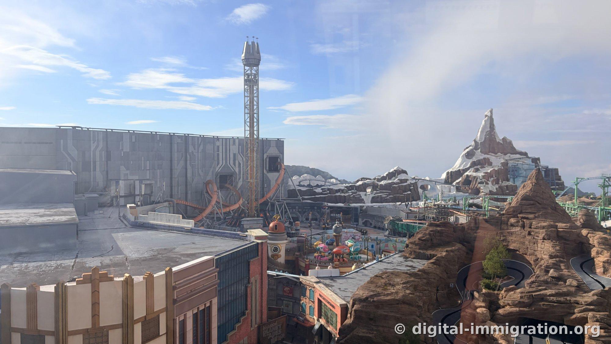 Drop tower and orange loop coaster in gray concrete area, red sandstone peak and snow mountain set to the right