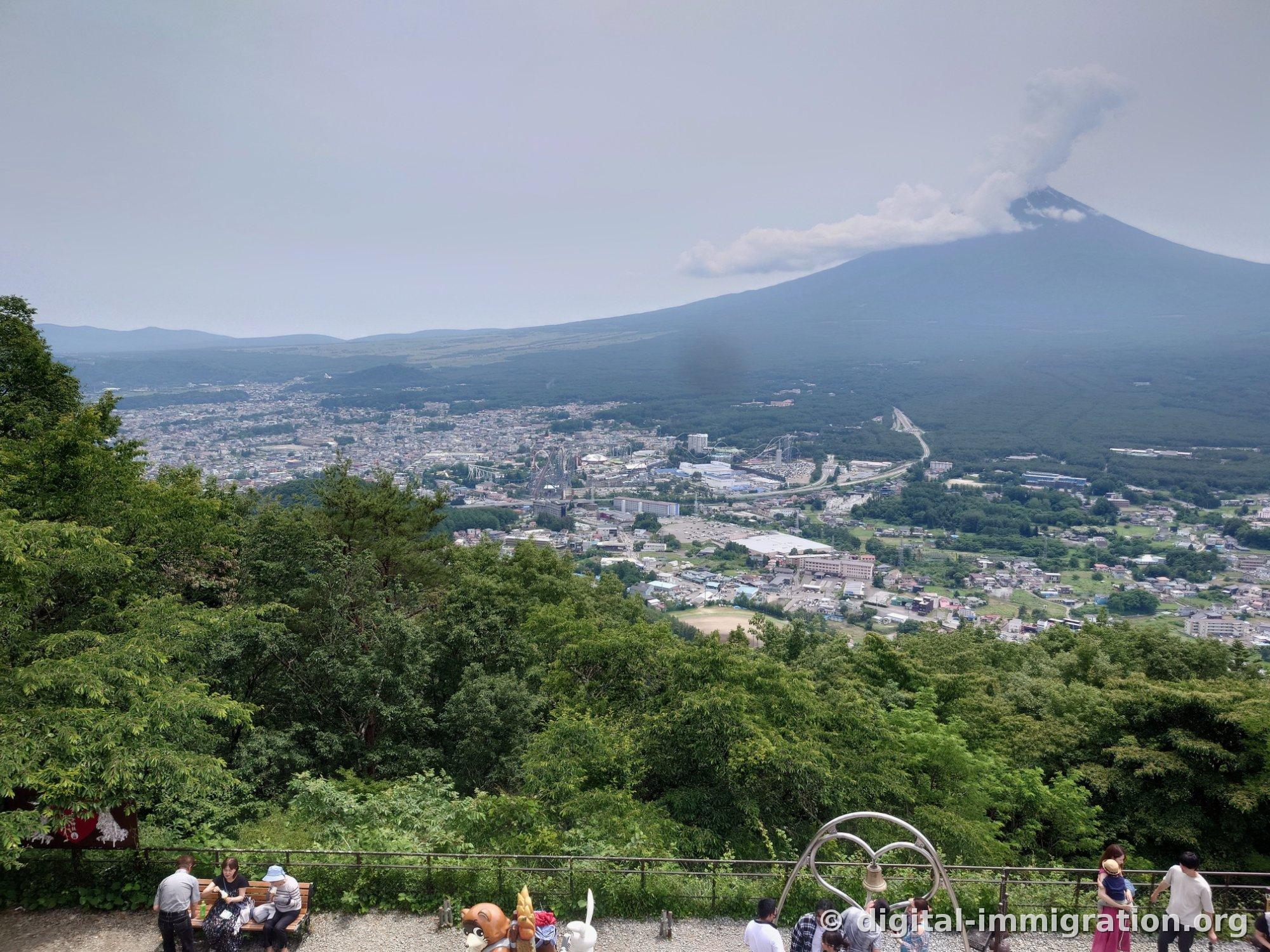 Clouds streaming from Mount Fuji's summit to the right, with Fujiyoshida town below and people sitting on benches at the observation deck in the foreground