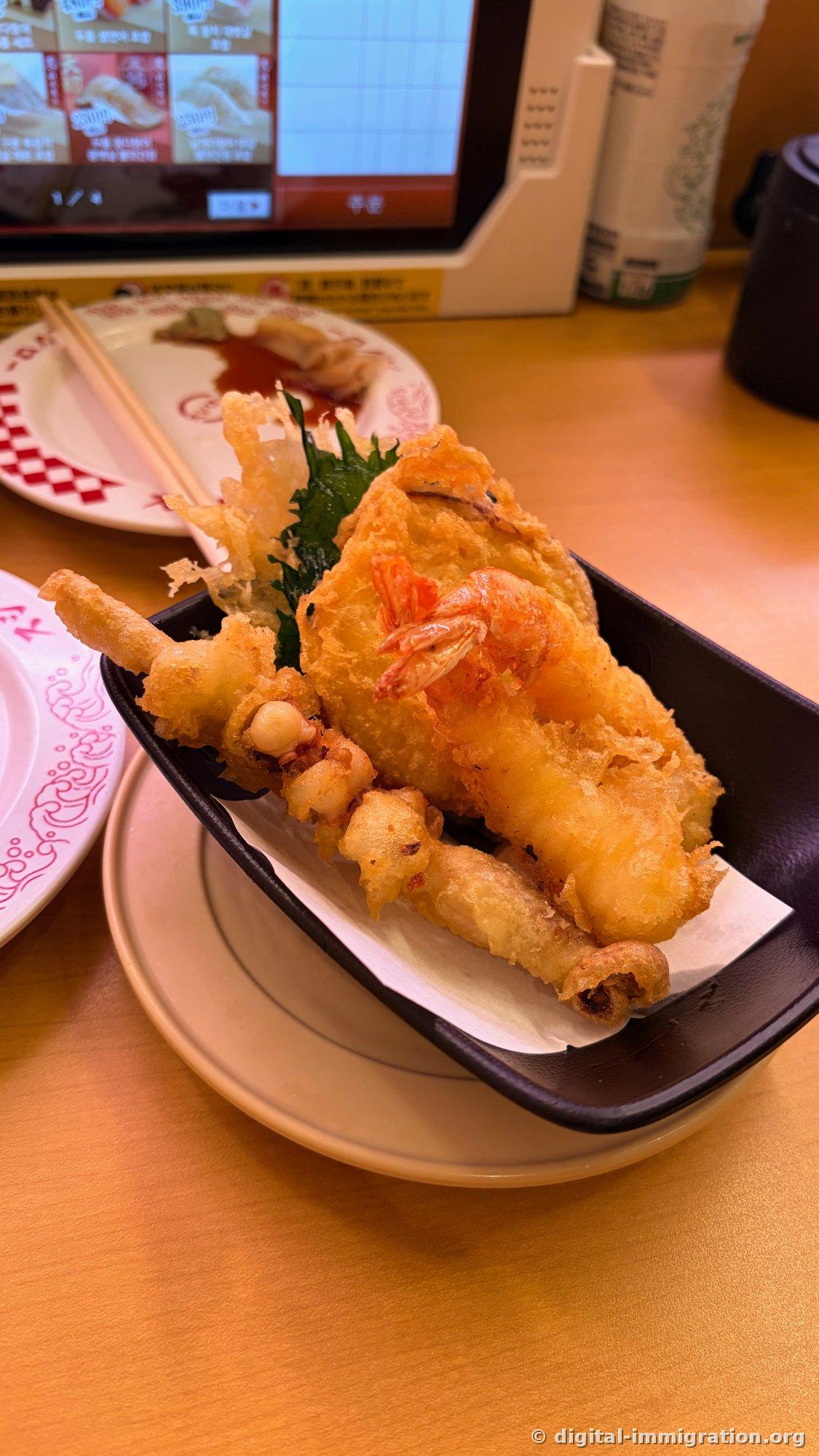 Un assortiment de crevettes, calmar et poisson blanc frits dans un bol en papier noir, avec une tablette de commande affichant le menu en coréen à l'arrière-plan