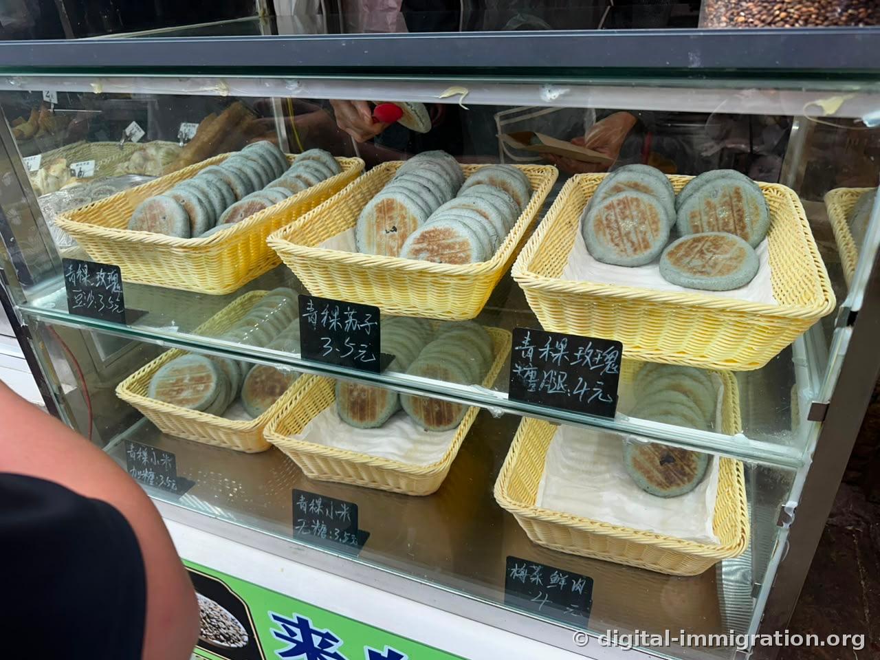 Pale green qingke cakes in yellow baskets inside a glass display case with a blackboard price sign reading '青稞 苏子 3.5元', '青稞 玫瑰 糖 4元'