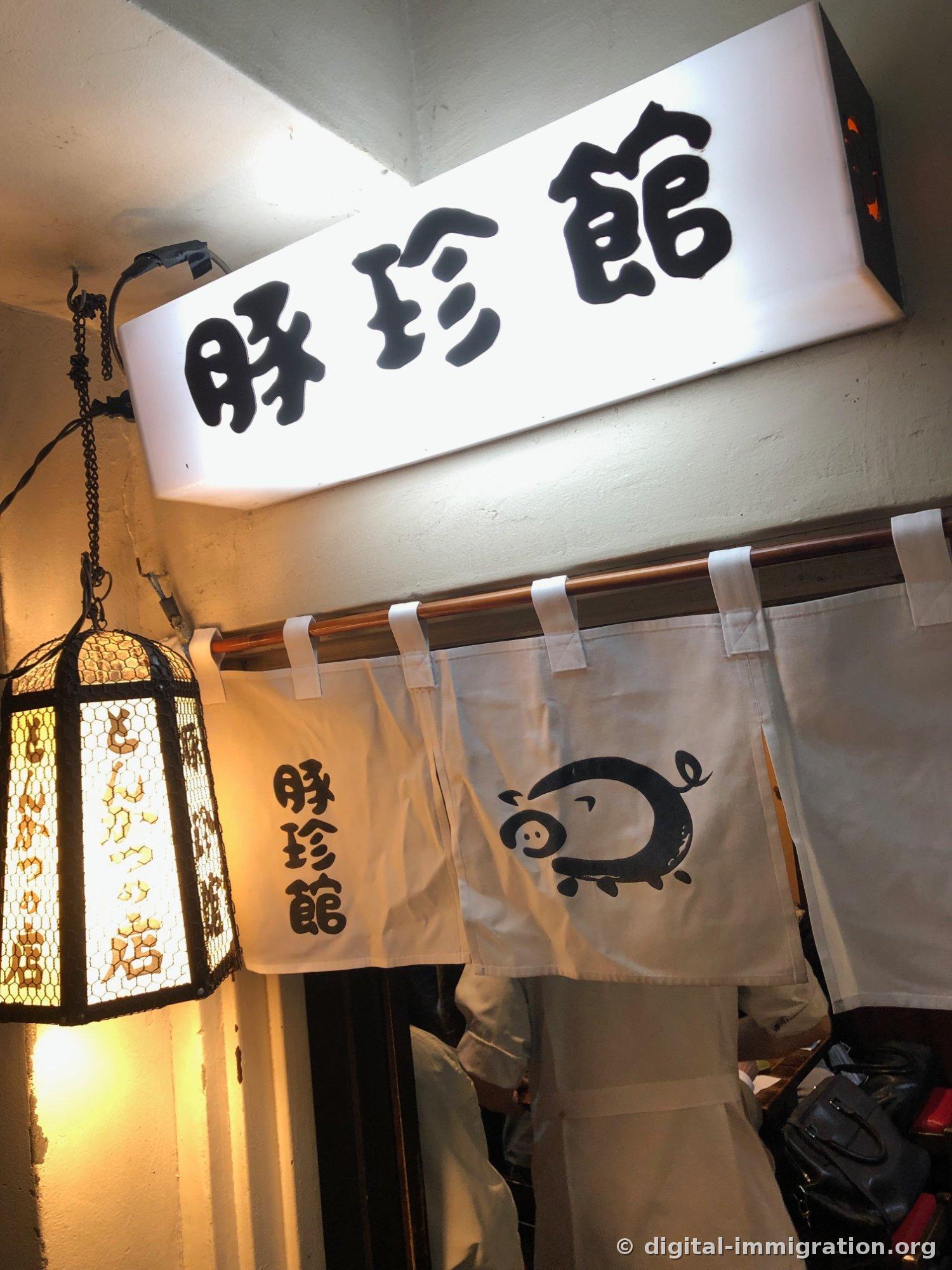 A shop entrance with large kanji characters '豚珍館' on a white LED sign, beneath which hangs a white noren curtain featuring a pig illustration, with a hexagonal lantern hanging to the left